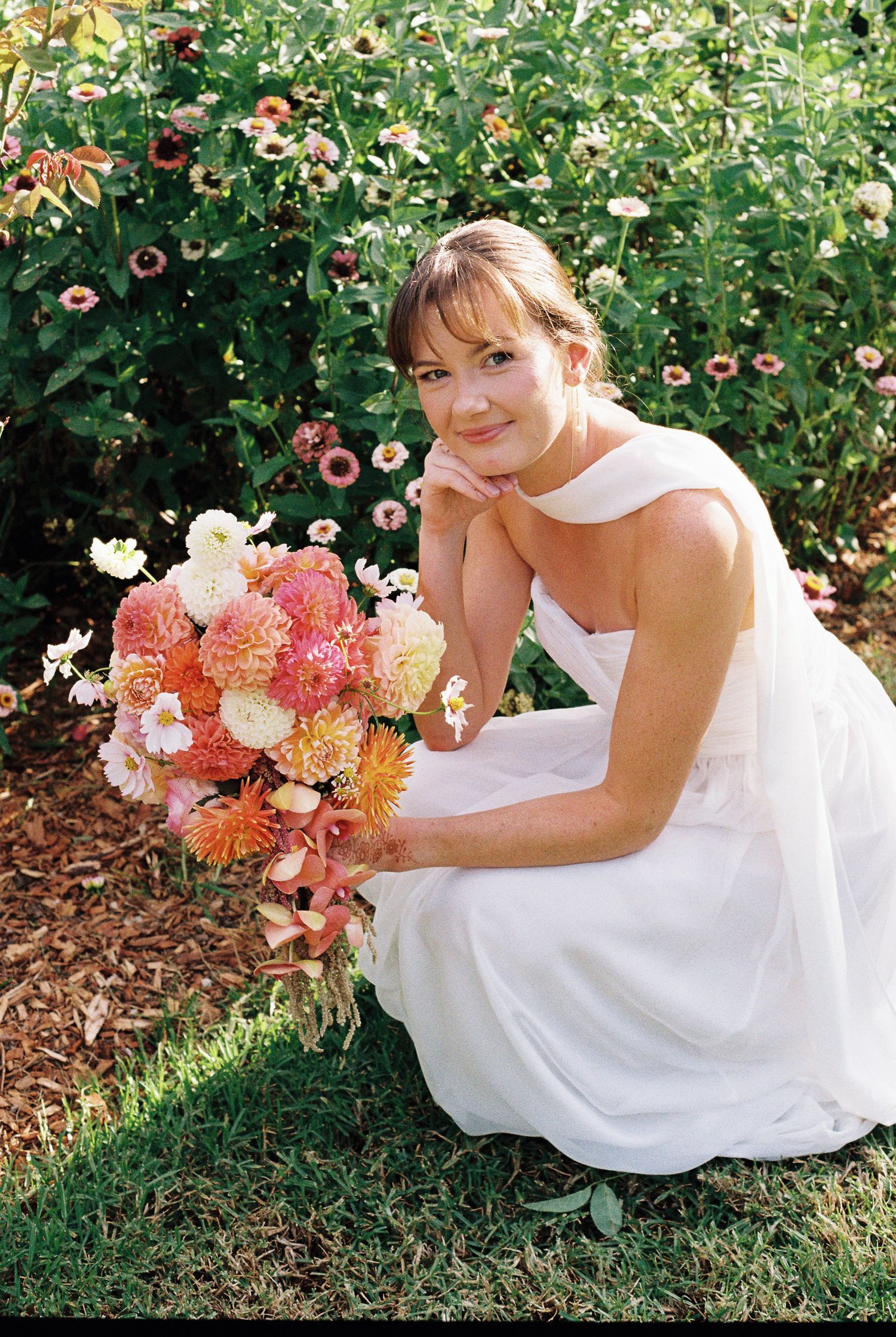 Smiling bride in a white one-shoulder dress kneels on grass holding a pink and orange zinnia bouquet in a sunny flower garden.