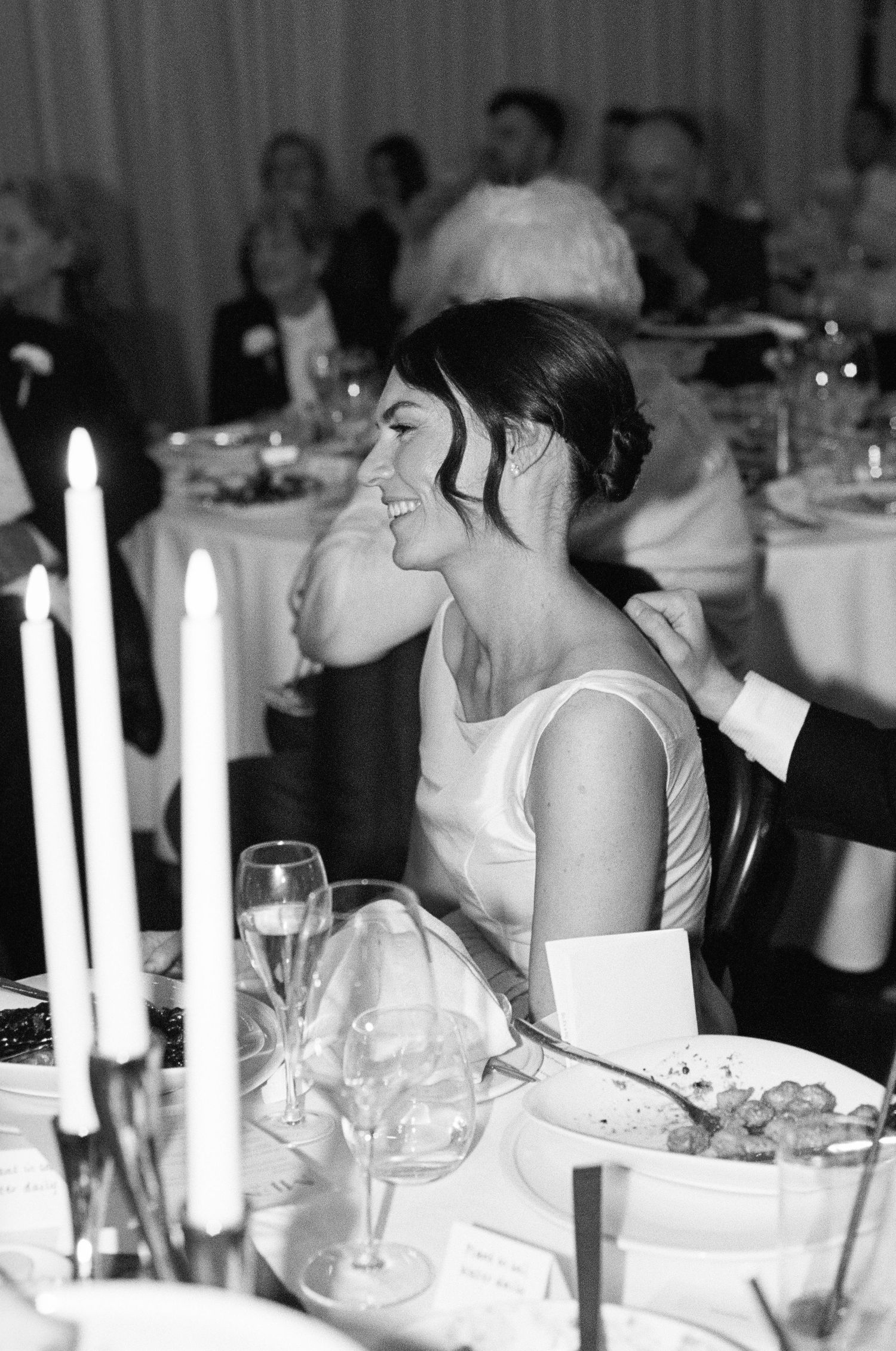 Smiling bride seated at wedding reception table with candles and glassware in black and white photo.