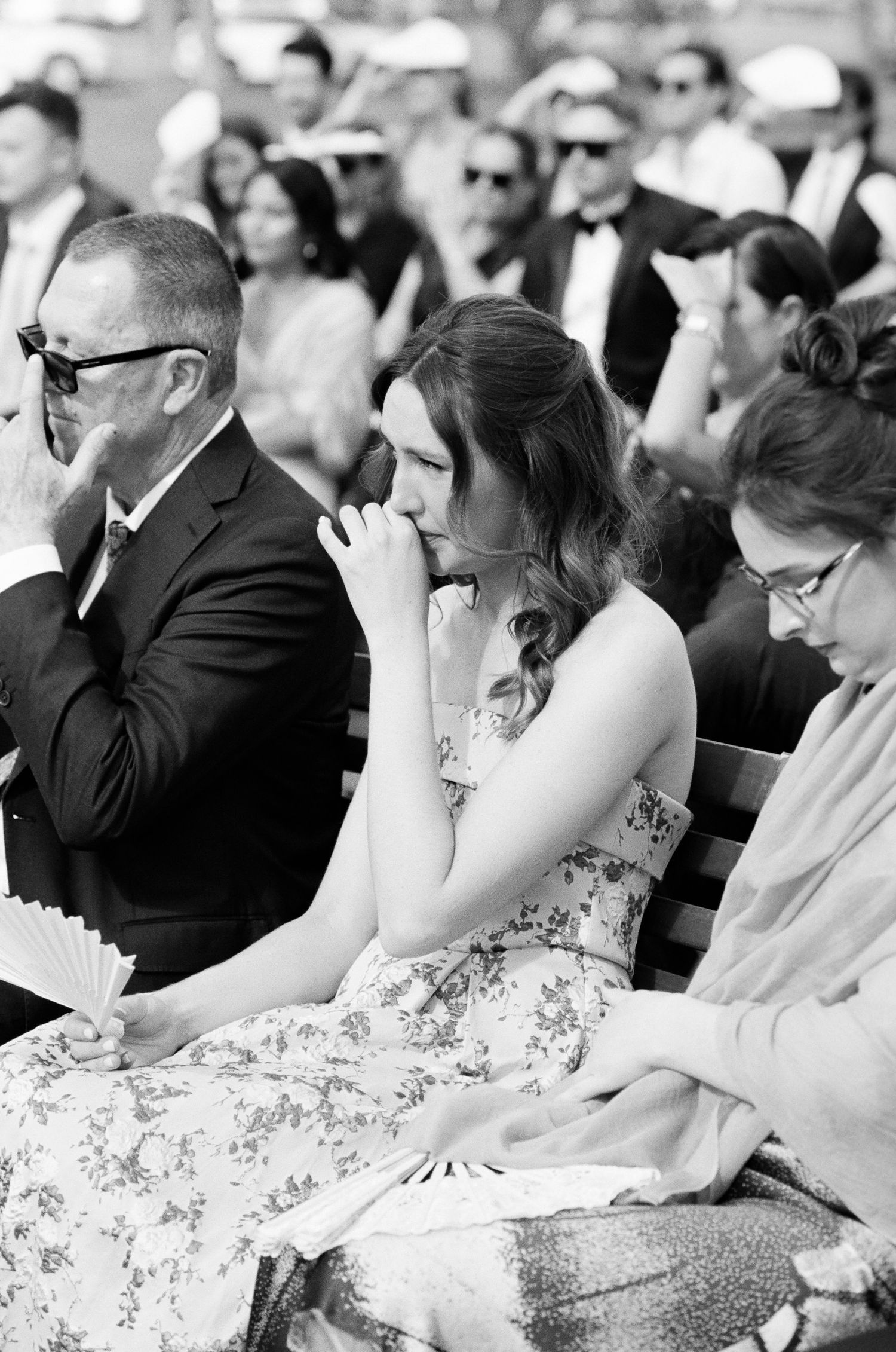Black and white photo of an emotional woman in a floral dress wiping tears while seated among wedding ceremony guests.
