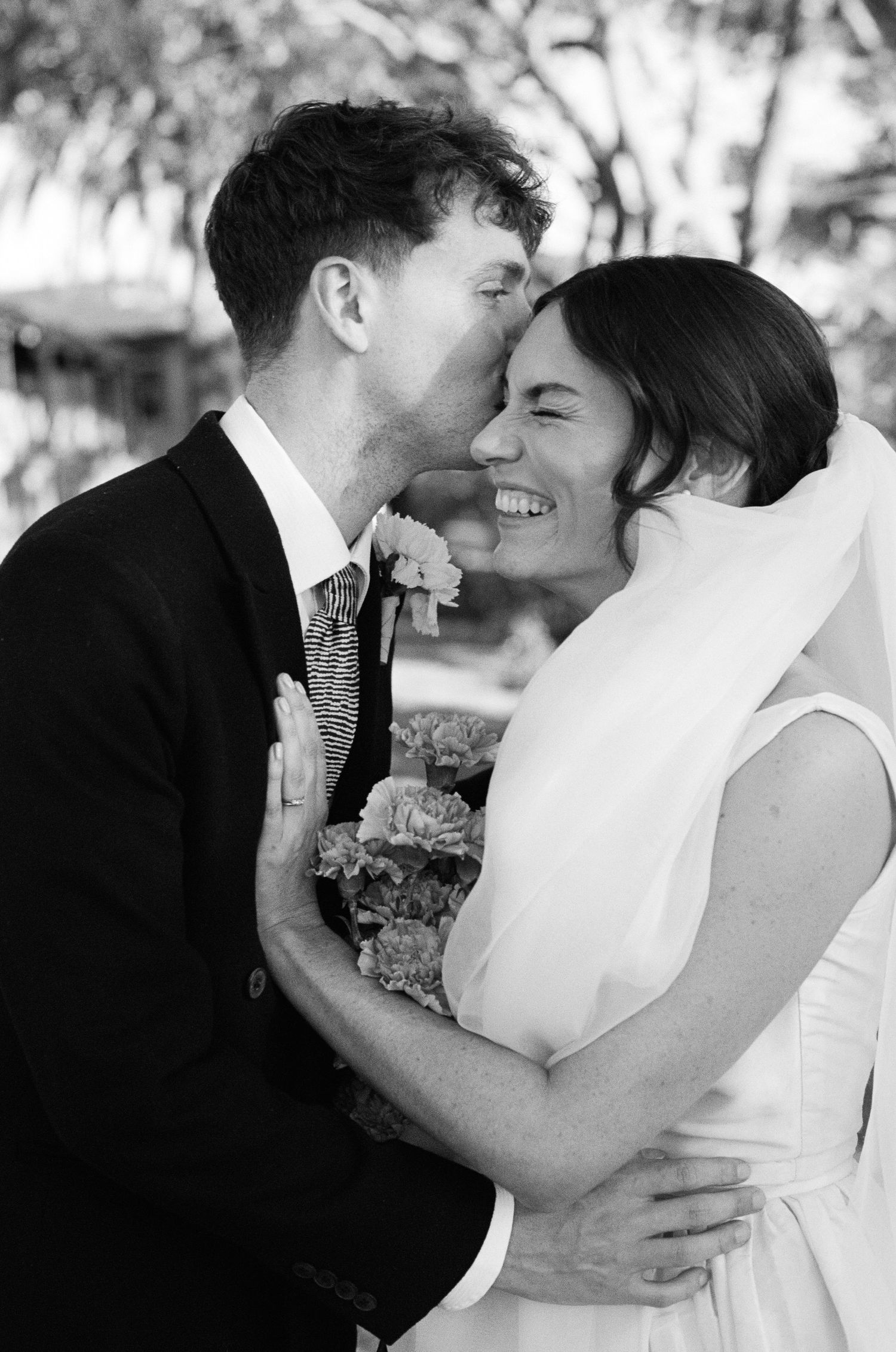 Black and white photo of groom kissing smiling bride on the cheek while holding a floral bouquet.