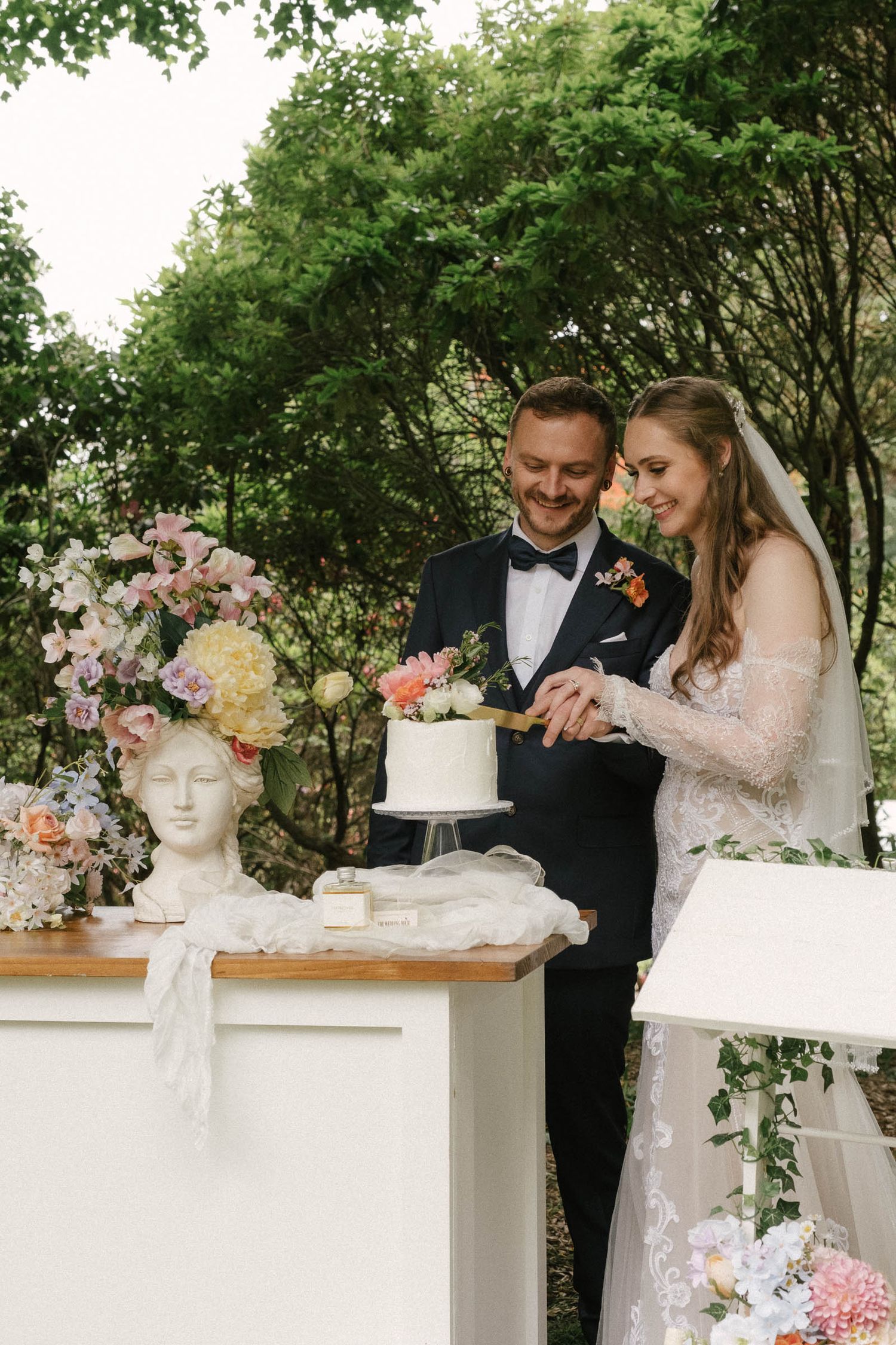 A romantic garden wedding scene with a couple cutting their elegant white cake surrounded by classical busts and floral decor.