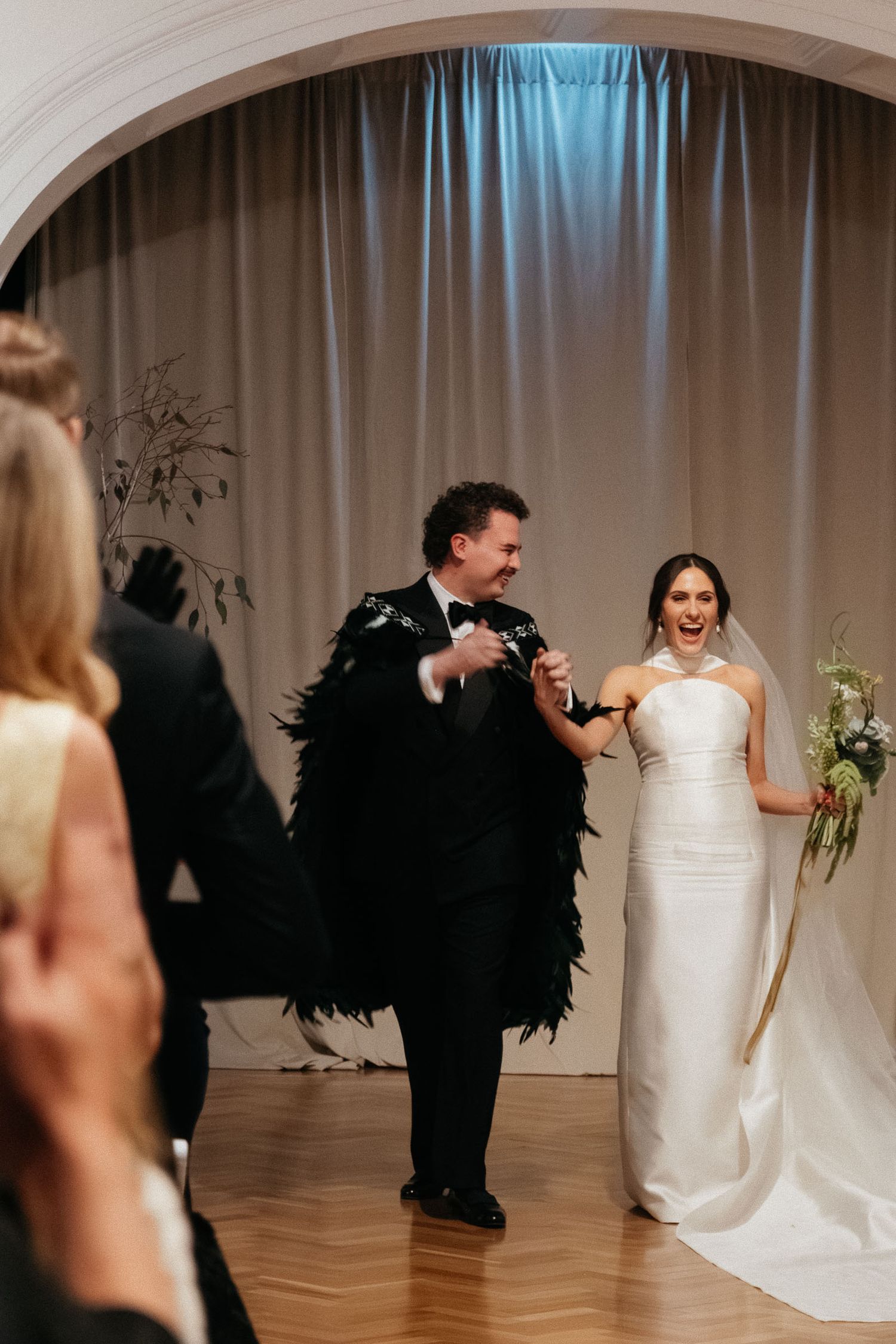 A couple in black and white attire dances playfully during their wedding reception on a wooden dance floor.