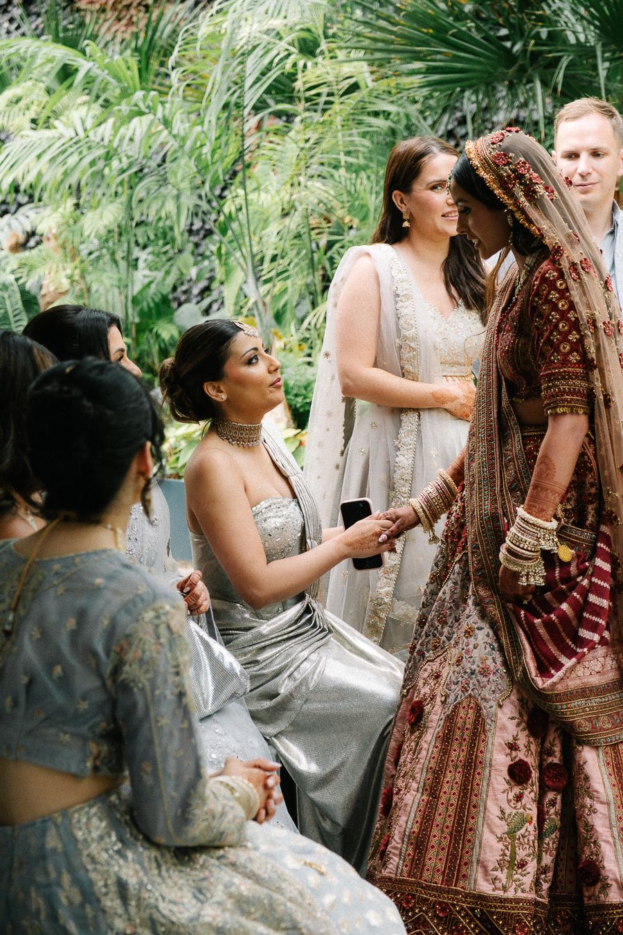 Wedding party members gather during a ceremony wearing elegant traditional Indian and Western formal attire in a garden setting.