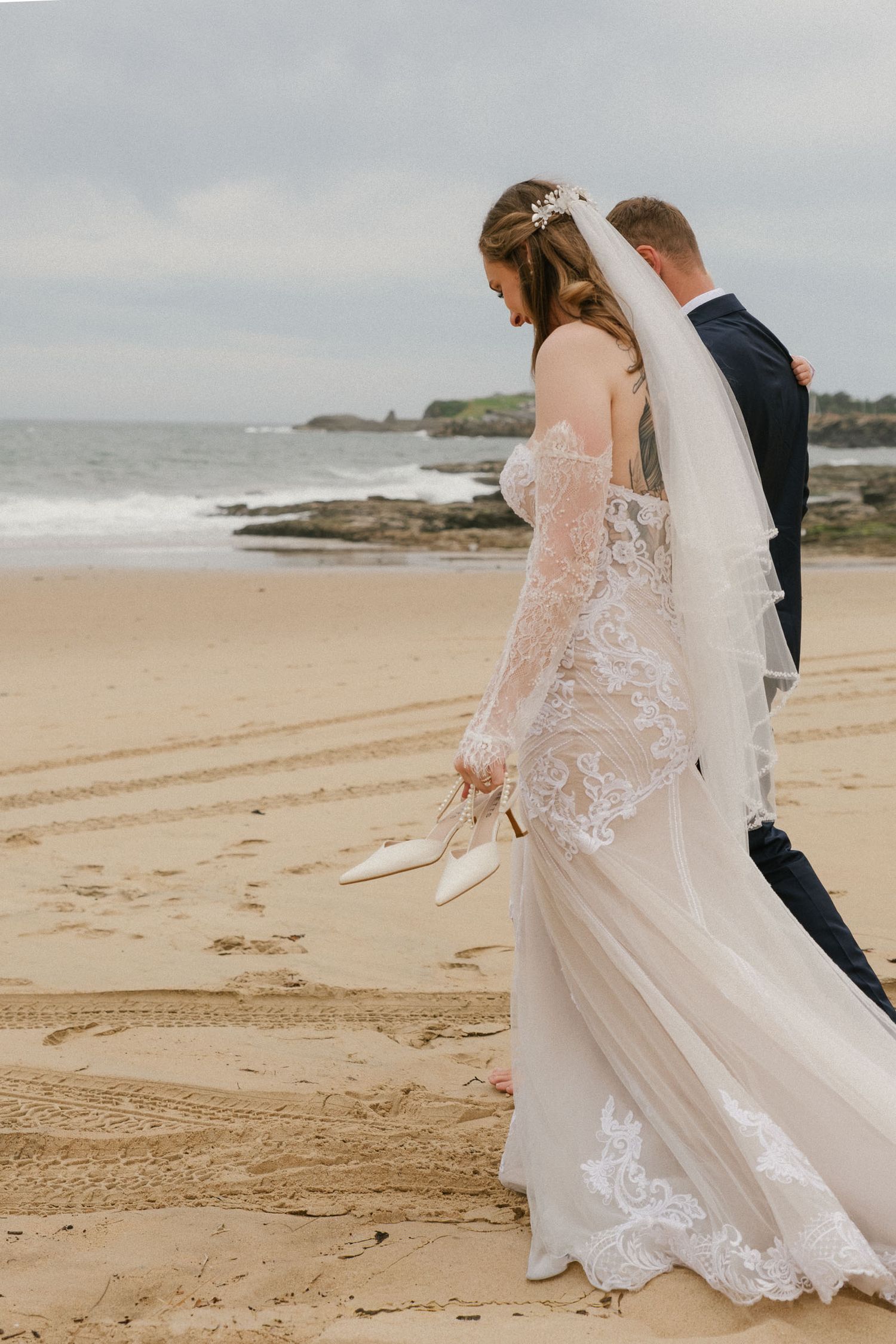 A couple walks along a sandy beach with the bride wearing a flowing lace wedding dress with long sleeves.