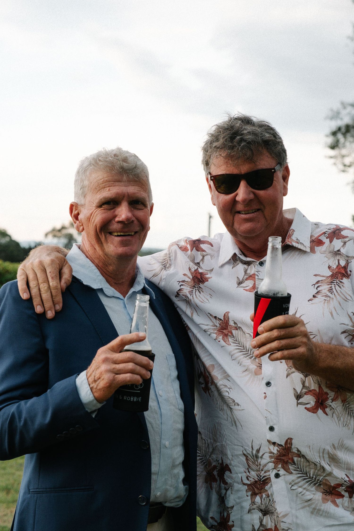 Two men share a laugh while embracing outdoors, one wearing a floral print shirt and the other in a navy blazer.