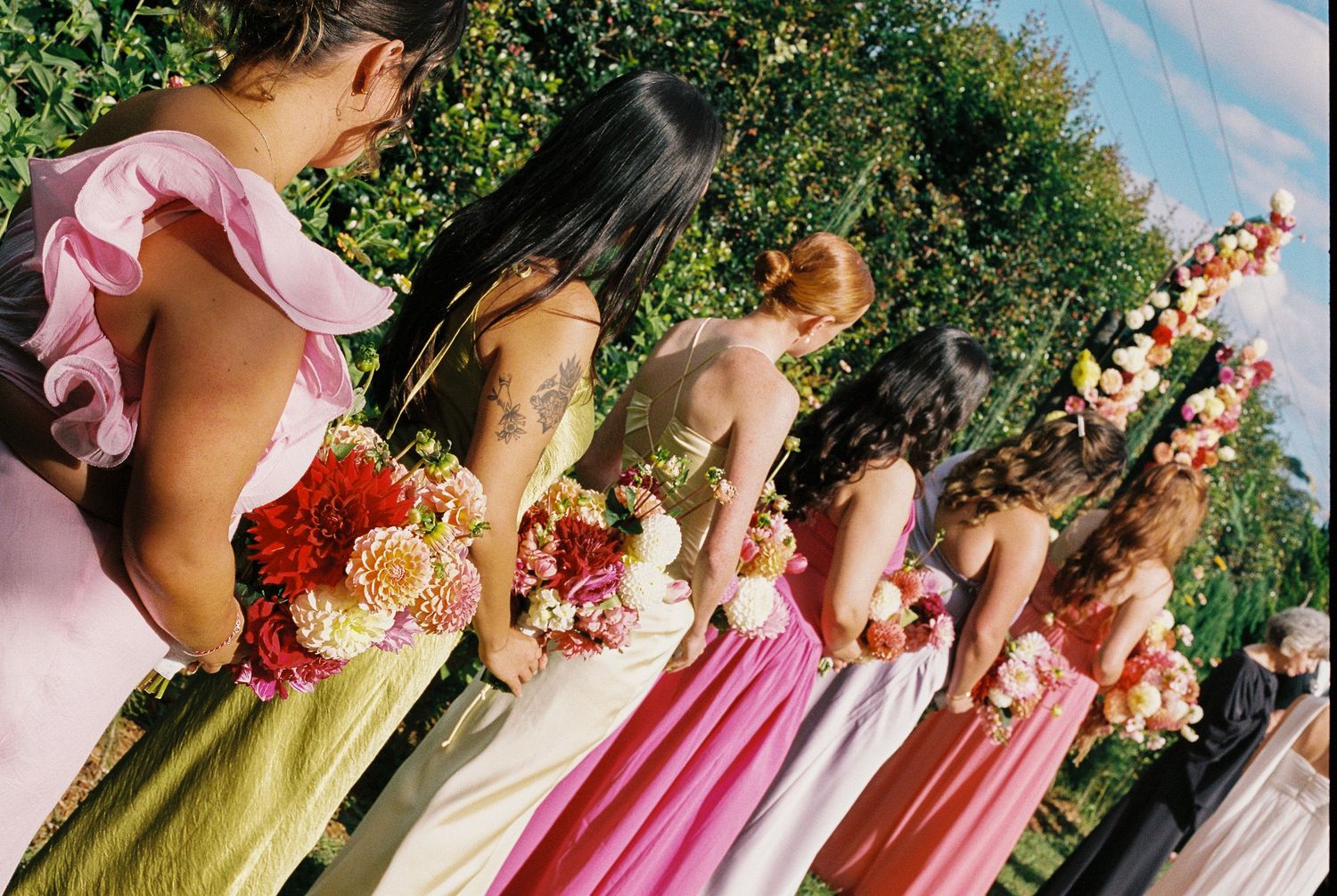 Bridesmaids in pink, coral and green dresses stand in a row outdoors holding colorful bouquets of orange, pink and white flowers.