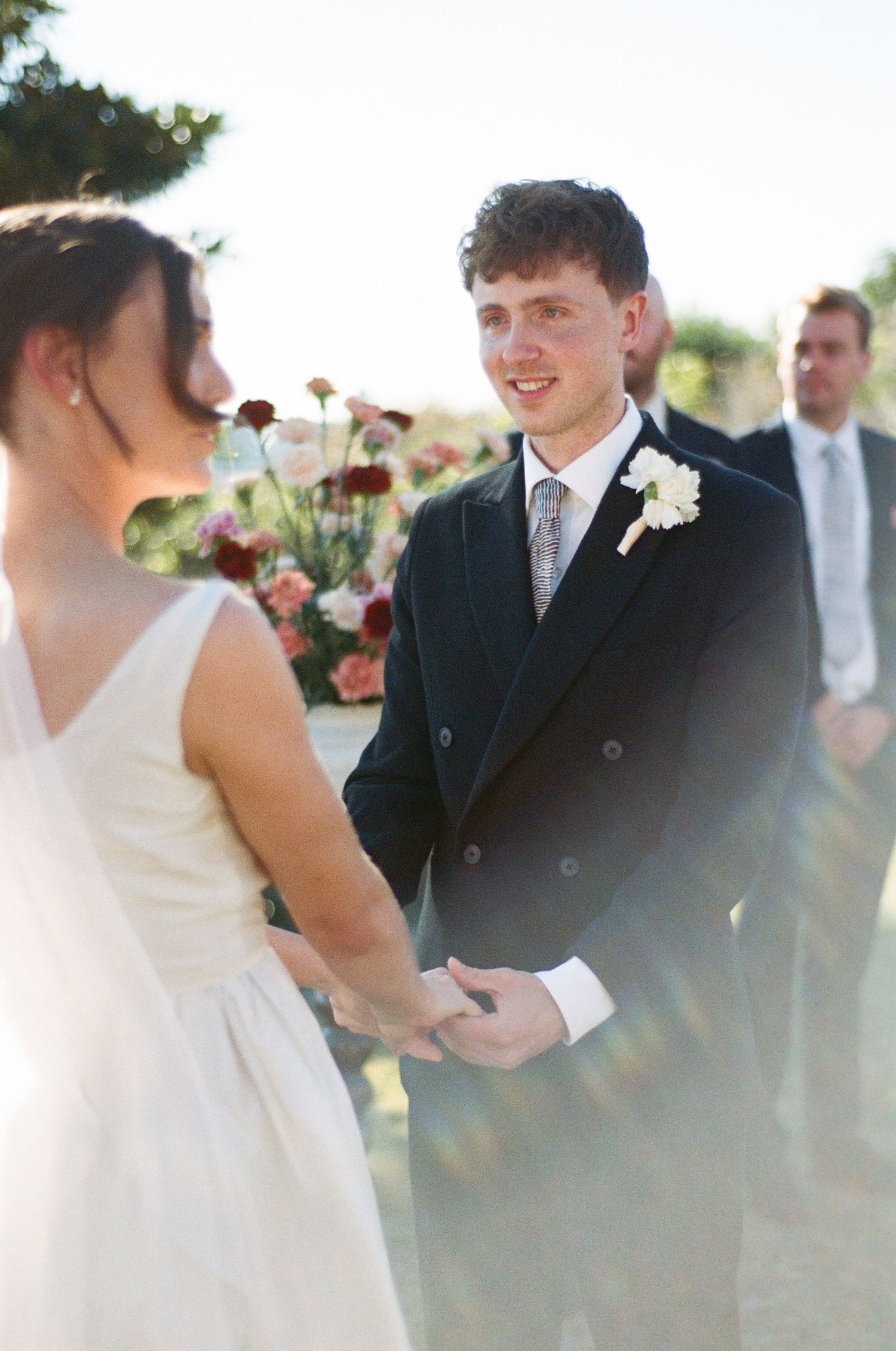 Groom in dark suit with boutonniere smiling at bride during outdoor wedding ceremony with floral arrangements.