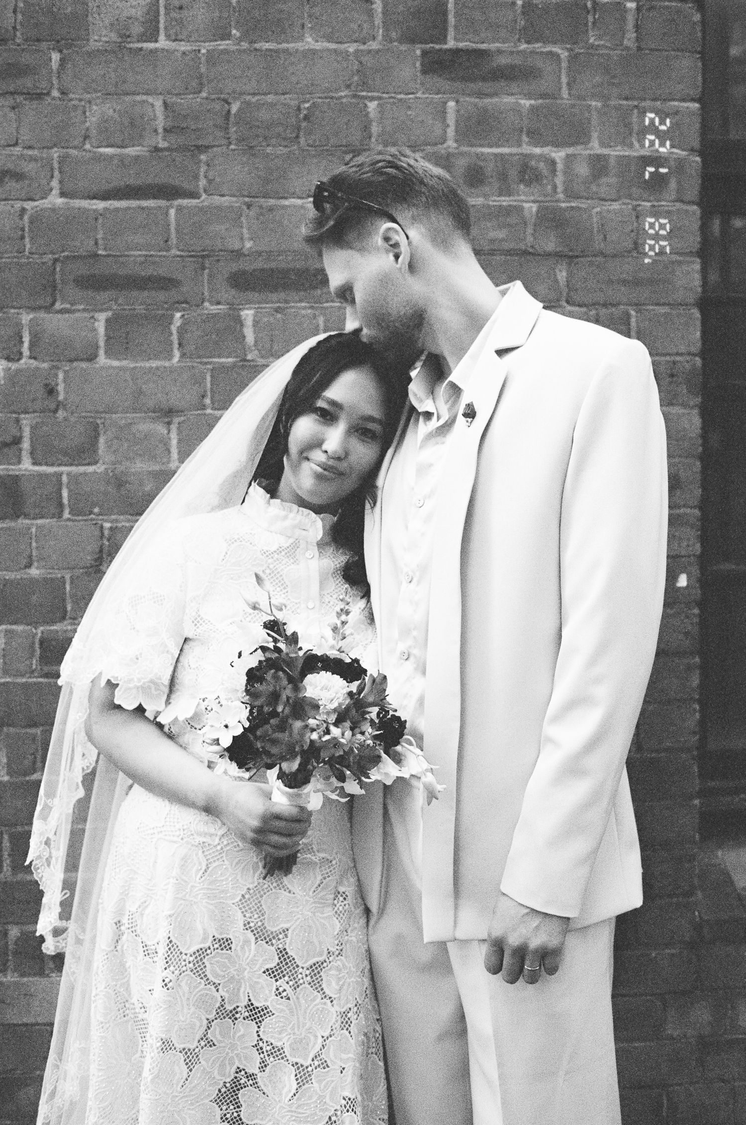 Black and white wedding portrait of a groom kissing his bride's forehead, she holds a bouquet in a lace dress and veil against a brick wall.