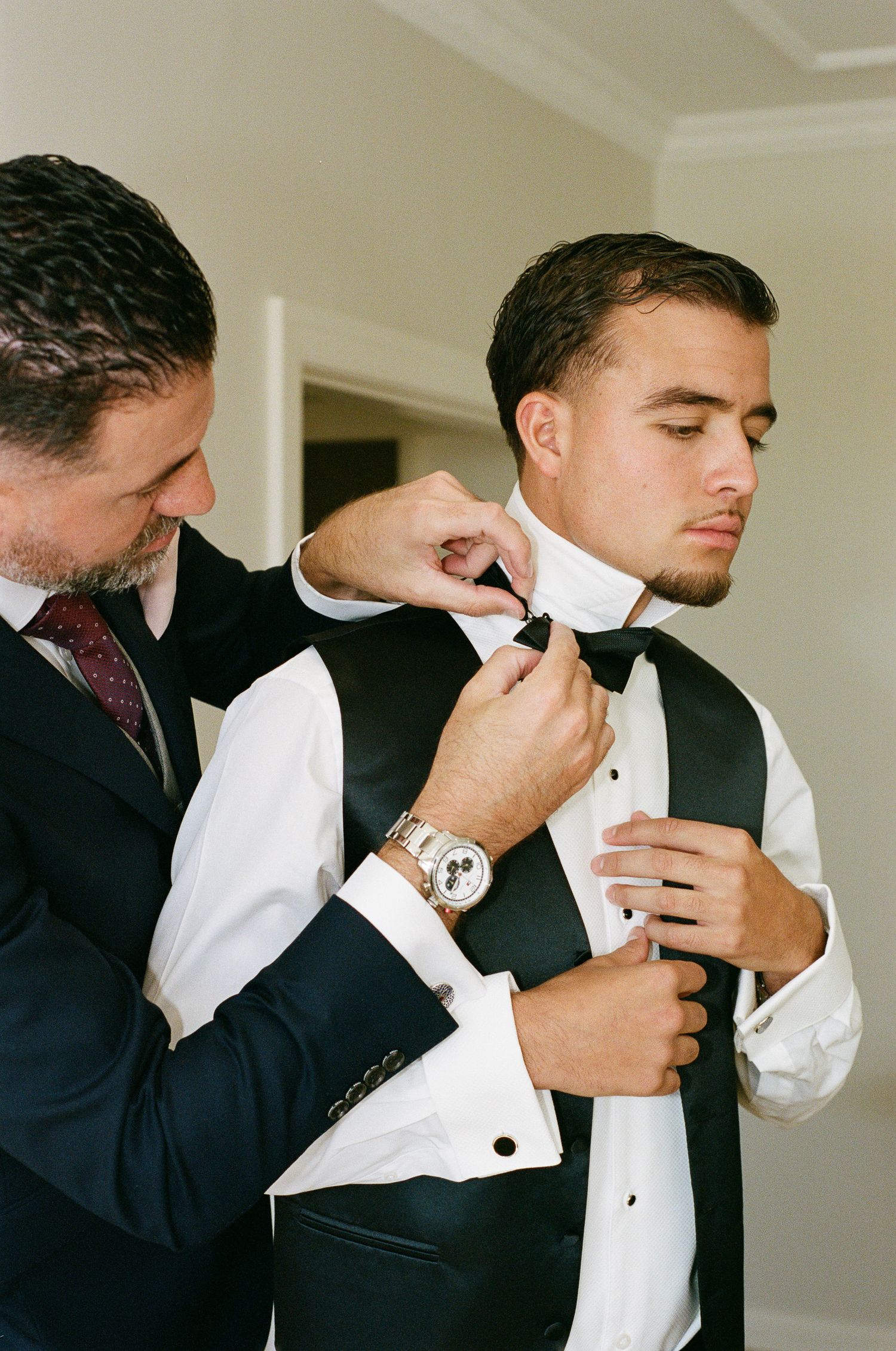 A man in a dark suit helps a groom in a white dress shirt and black vest adjust his bow tie while getting ready for the wedding.