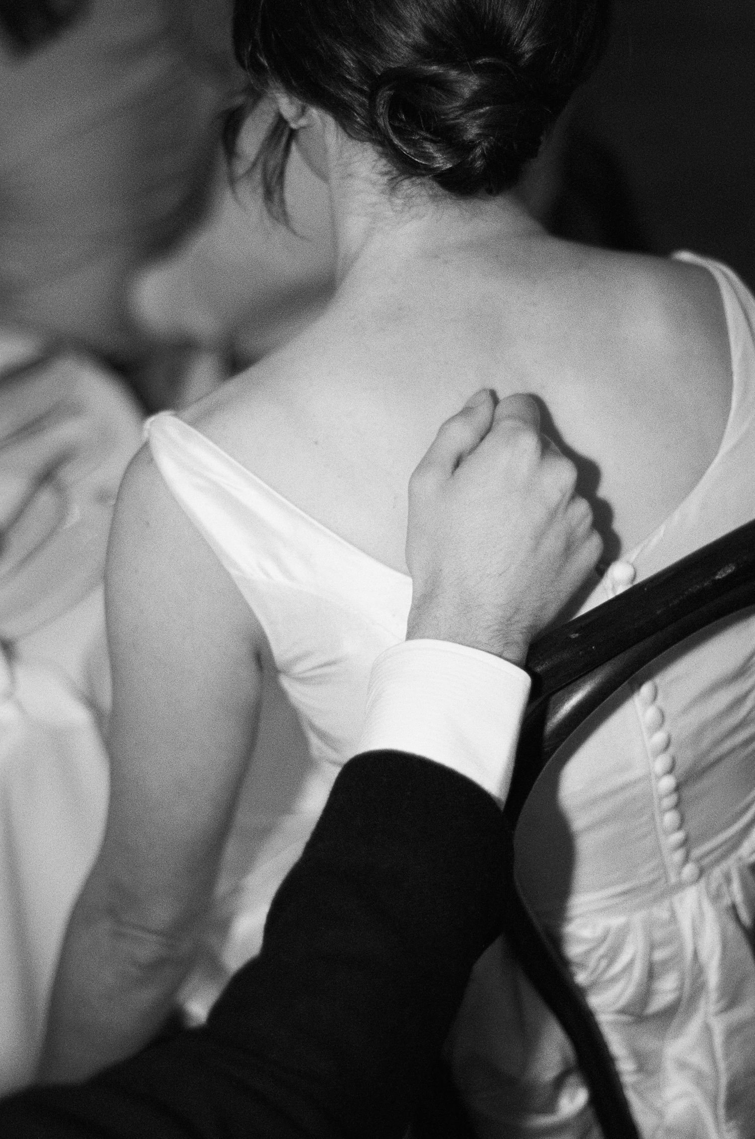 Black and white photo of a groom's hand resting on the back of a bride in a white dress with buttons and an updo.