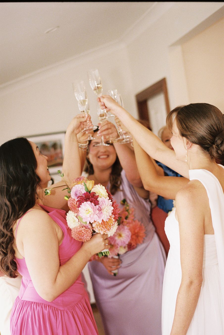 Bridesmaids in pink and lavender dresses toasting champagne flutes with bride holding colorful floral bouquet indoors.