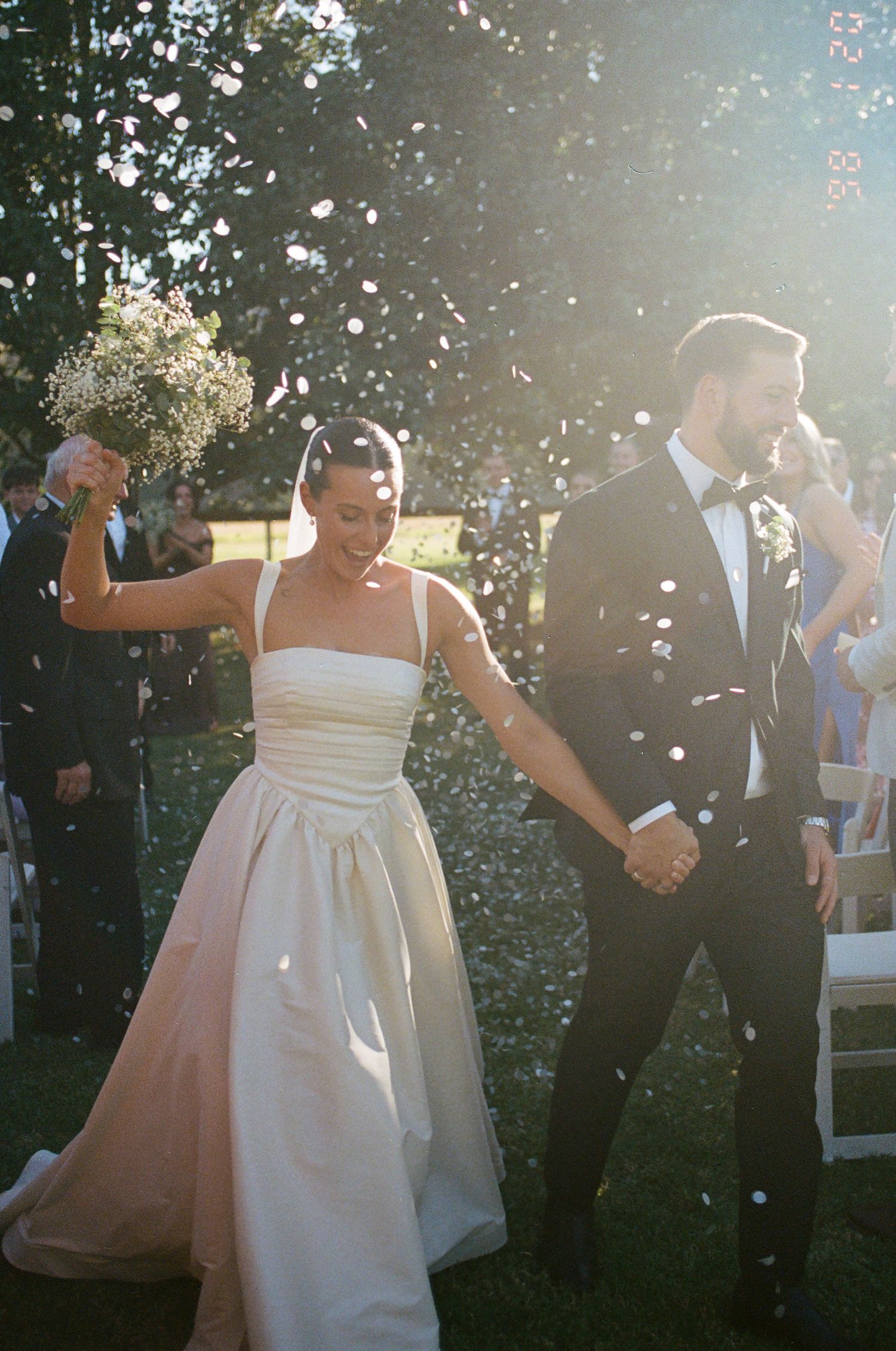 Smiling bride in a strapless white gown holds hands with her groom in a black tuxedo as confetti falls during an outdoor wedding exit.