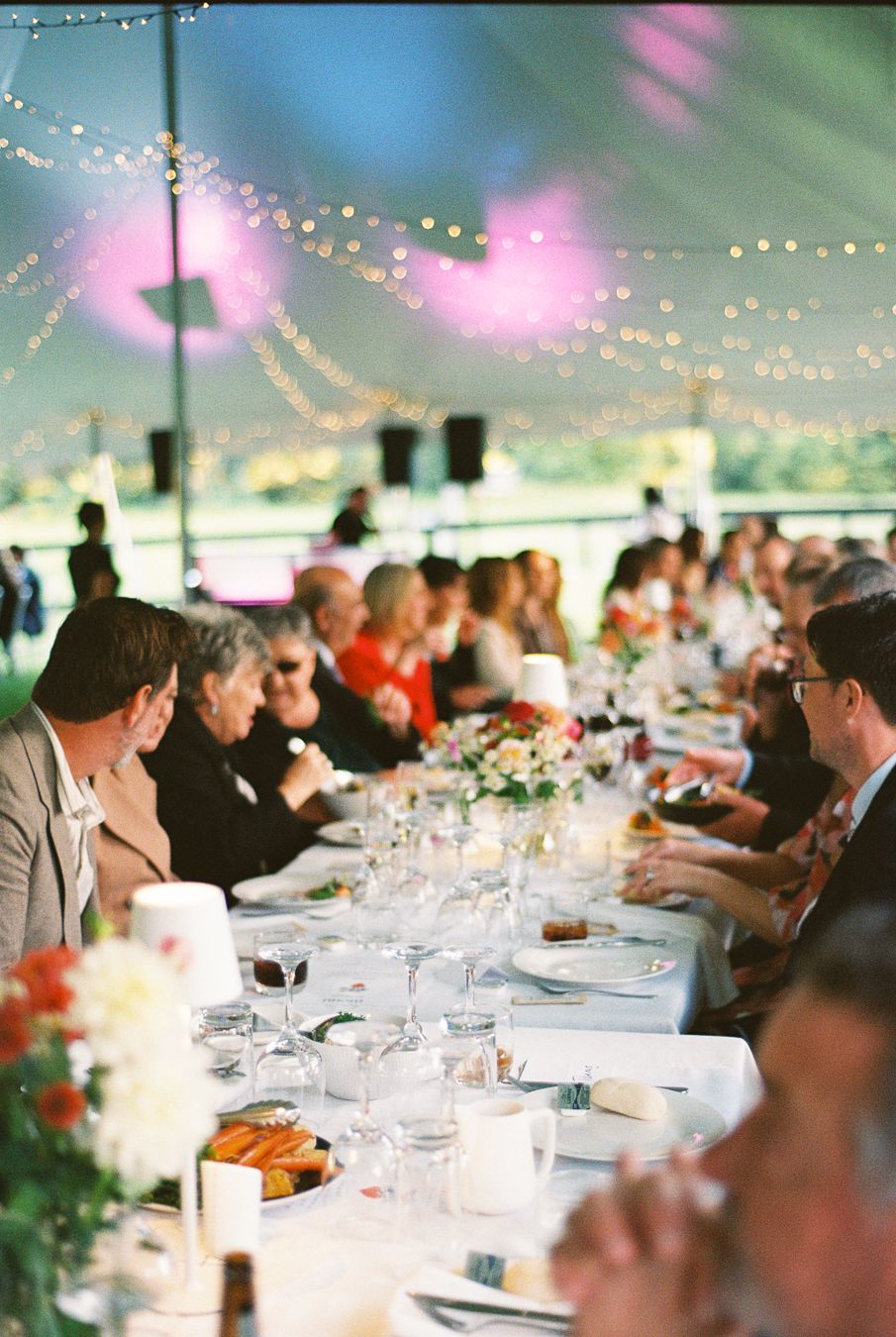 Elegant wedding reception under a tent with string lights, guests seated at long tables with floral centerpieces and glassware.