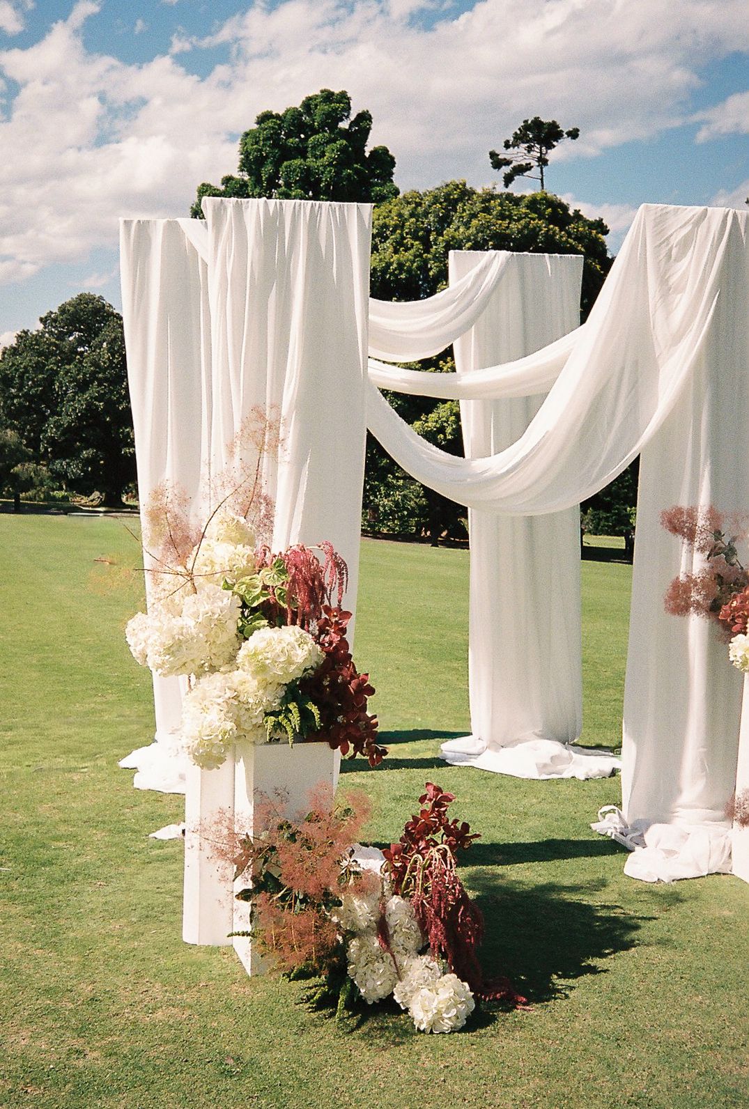 Elegant outdoor wedding ceremony setup with white fabric draping between pillars and floral arrangements on a green lawn.