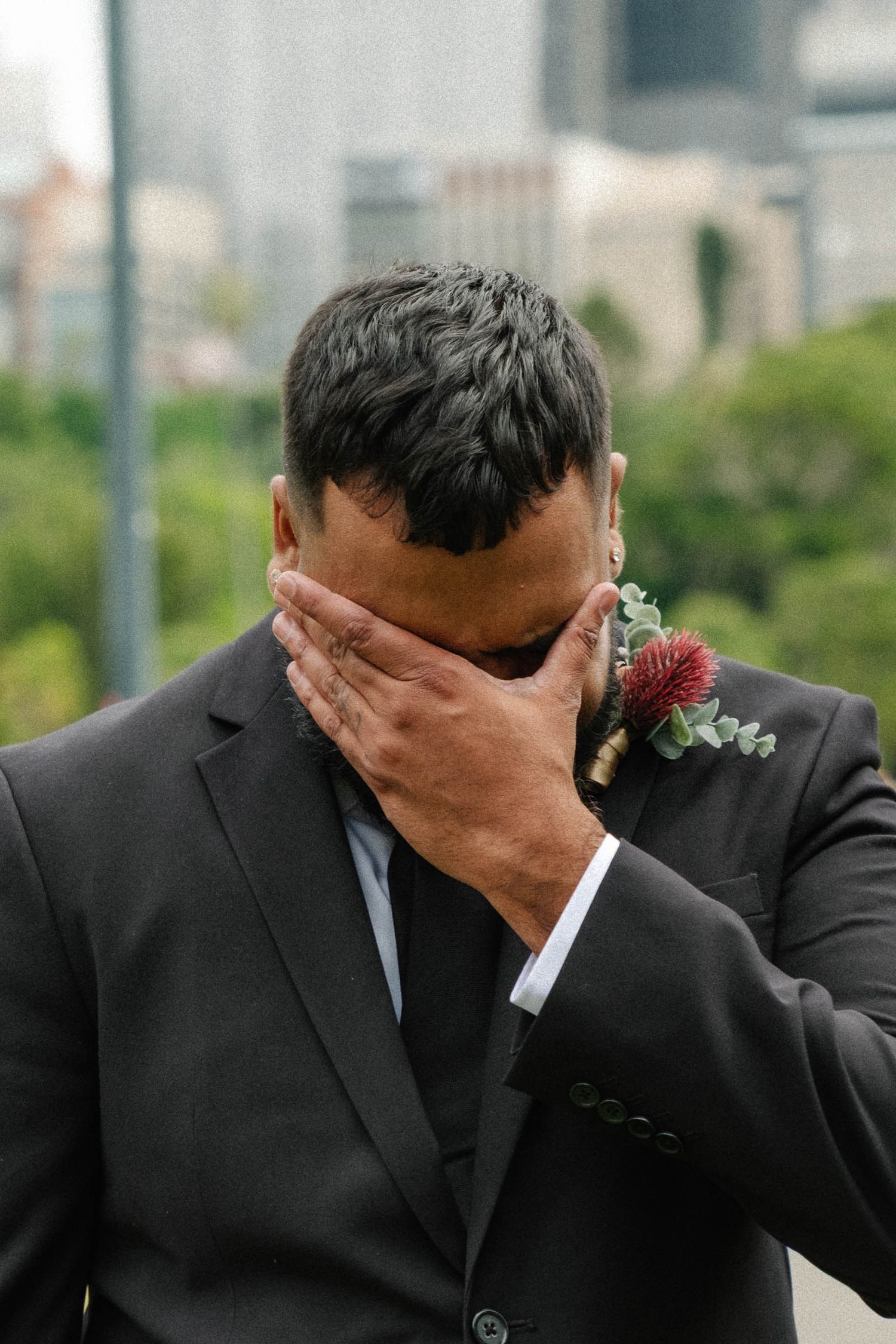 A person in a dark suit covers their face with their hand while wearing a red boutonniere.