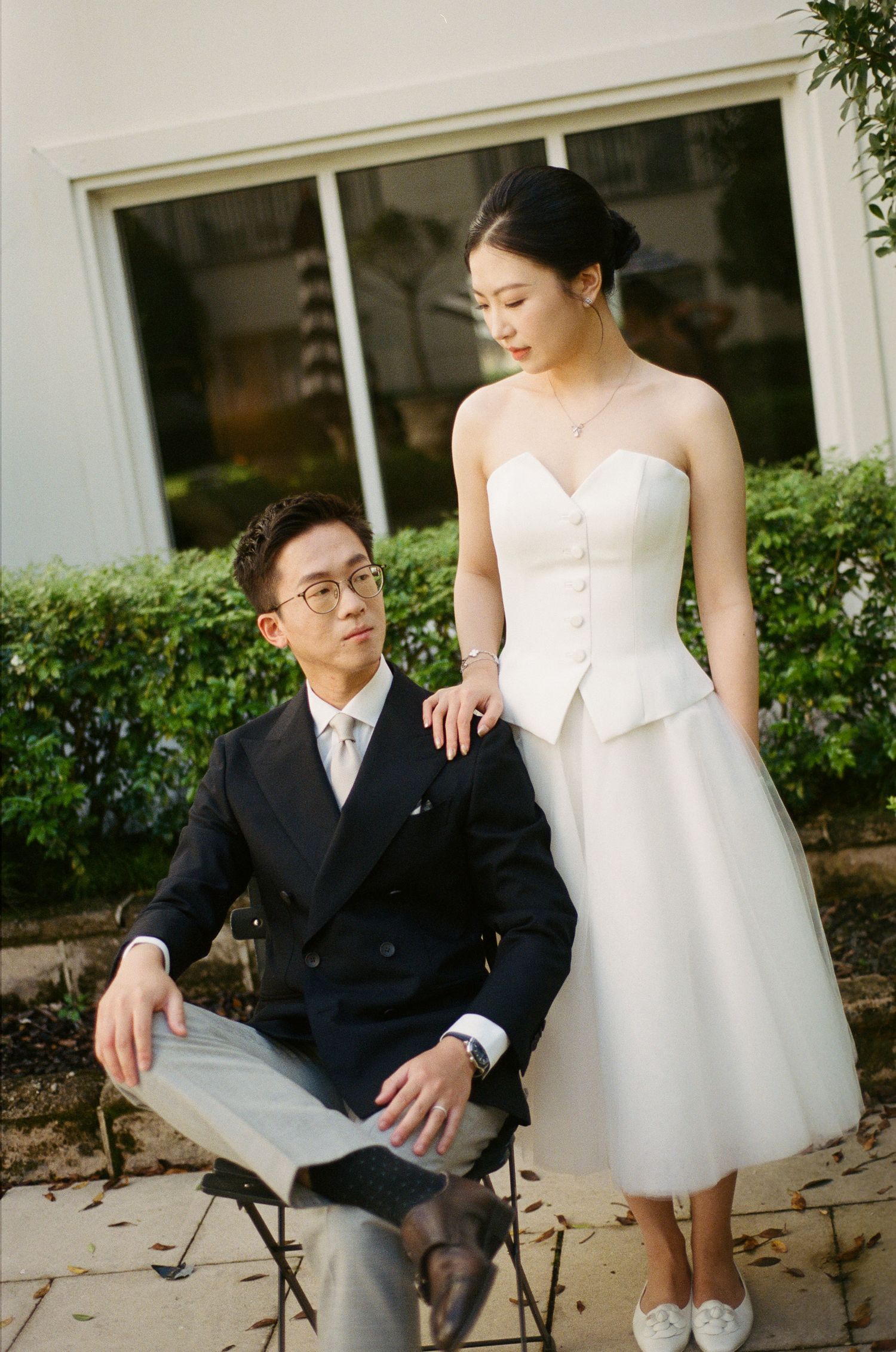 A man in a navy double-breasted suit and glasses sits on a folding stool while a woman in a white strapless dress stands beside him.
