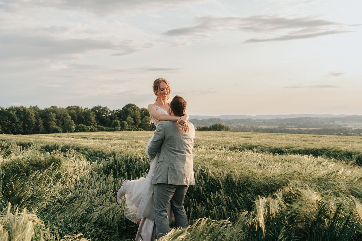 A couple embraces while standing in a golden wheat field at sunset with rolling hills in the background.