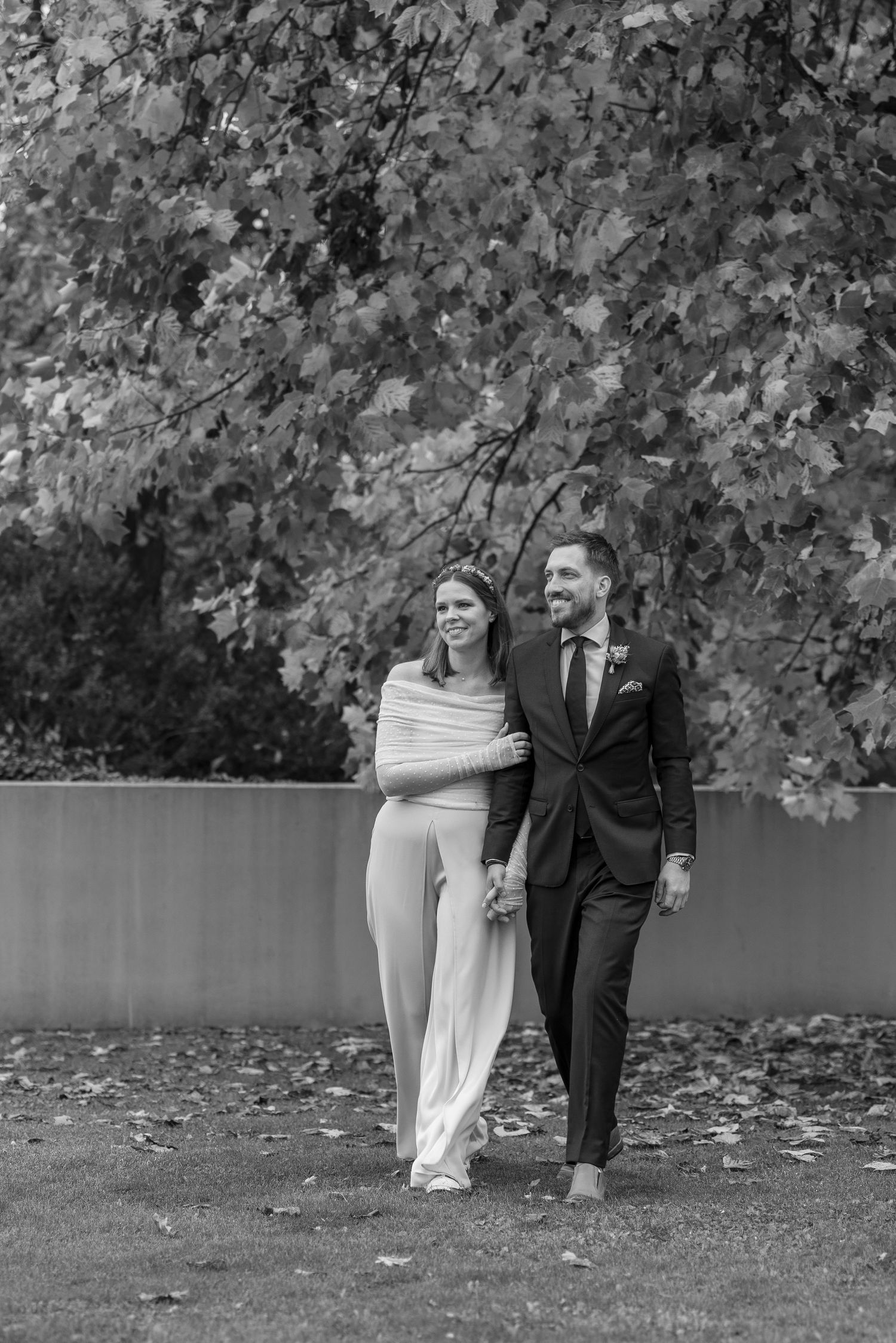 A couple in formal attire poses together by a lake surrounded by autumn trees in black and white.