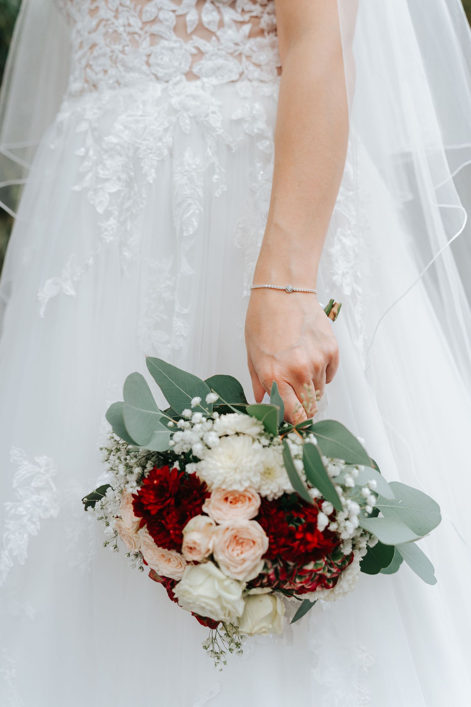 Close-up view of a bridal bouquet with burgundy and blush roses, white flowers and eucalyptus against a white dress.