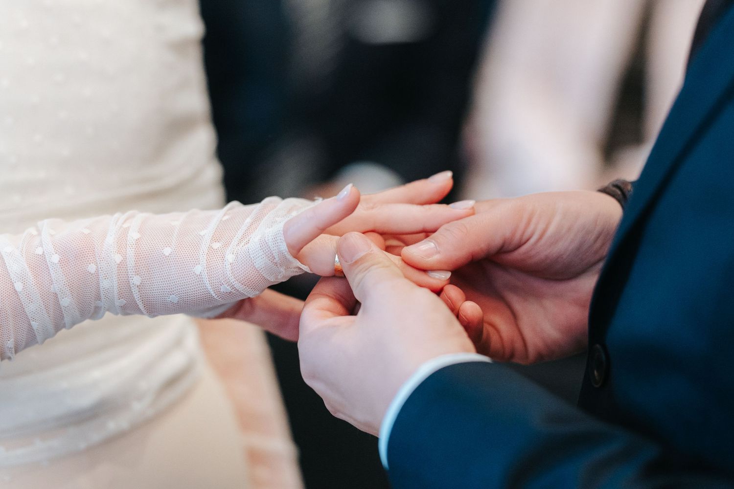 Close-up view of hands exchanging wedding rings during a ceremony with a navy suit and white dress visible.