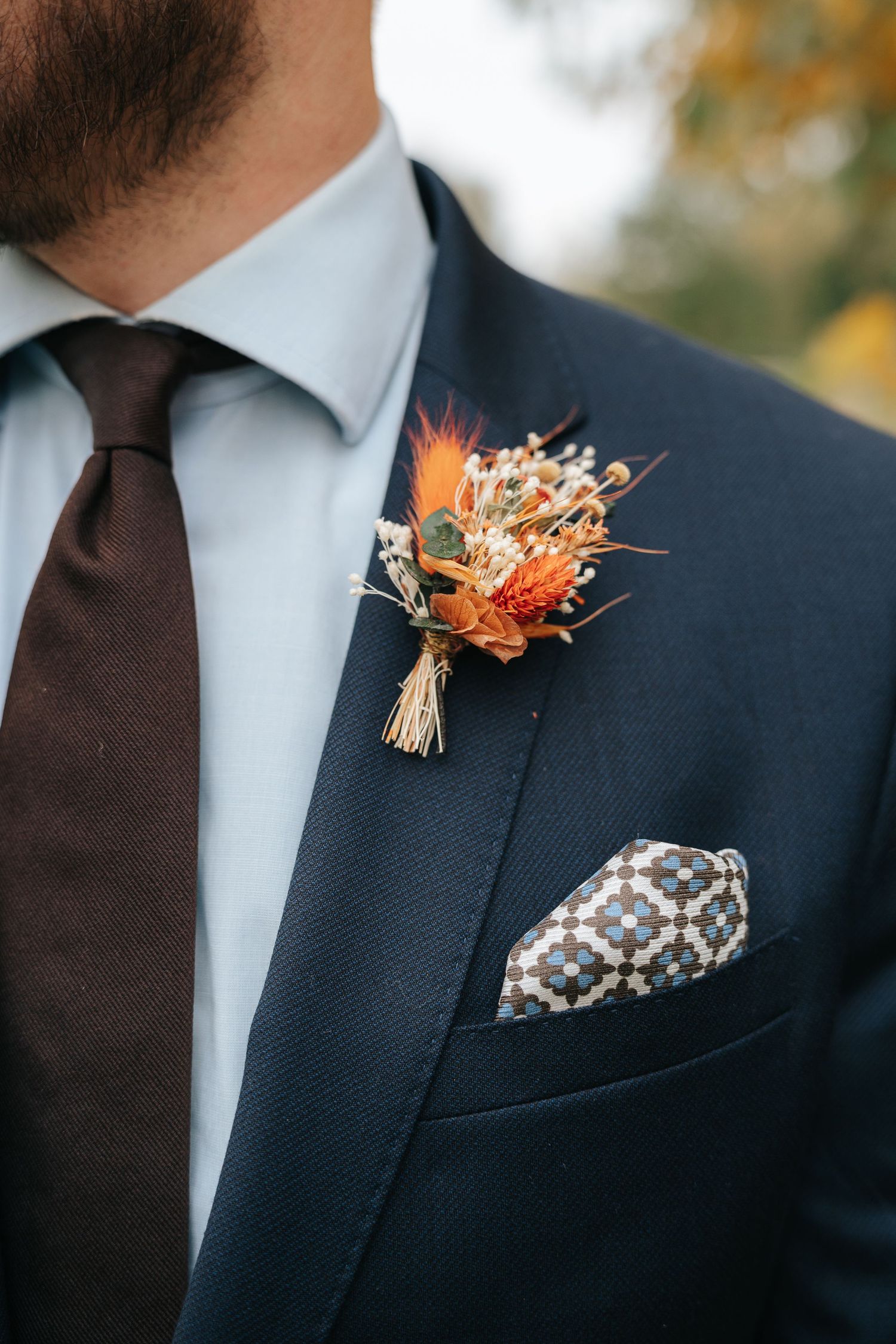 Close-up of navy suit with brown tie, patterned pocket square and orange dried flower boutonniere.