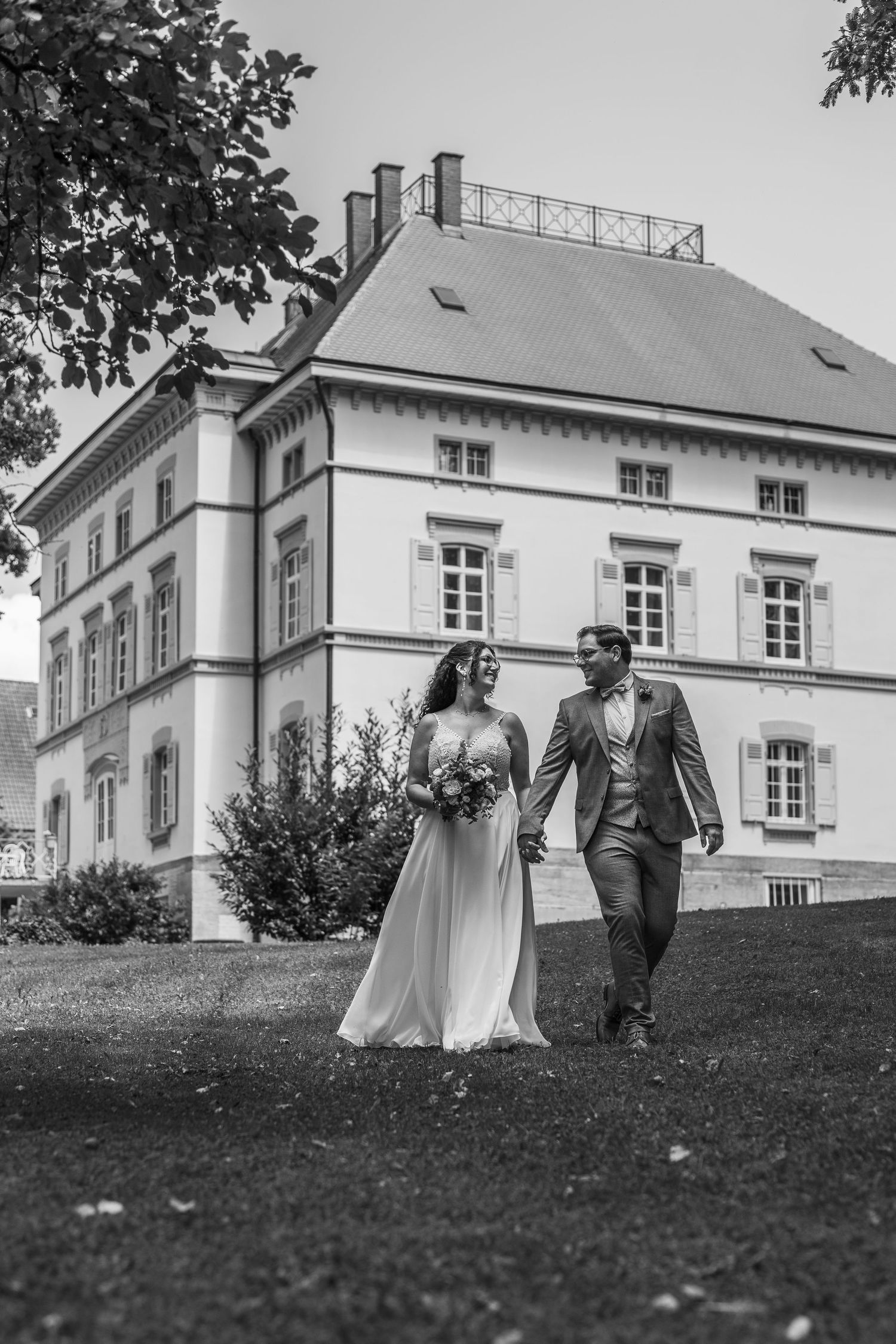Black and white wedding photos show a couple walking on a grassy hill near an elegant white mansion.
