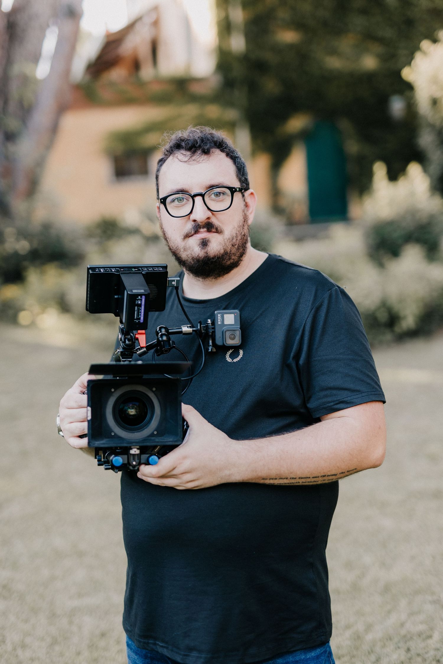 Bearded man with glasses holding a professional cinema camera outdoors in a garden setting.