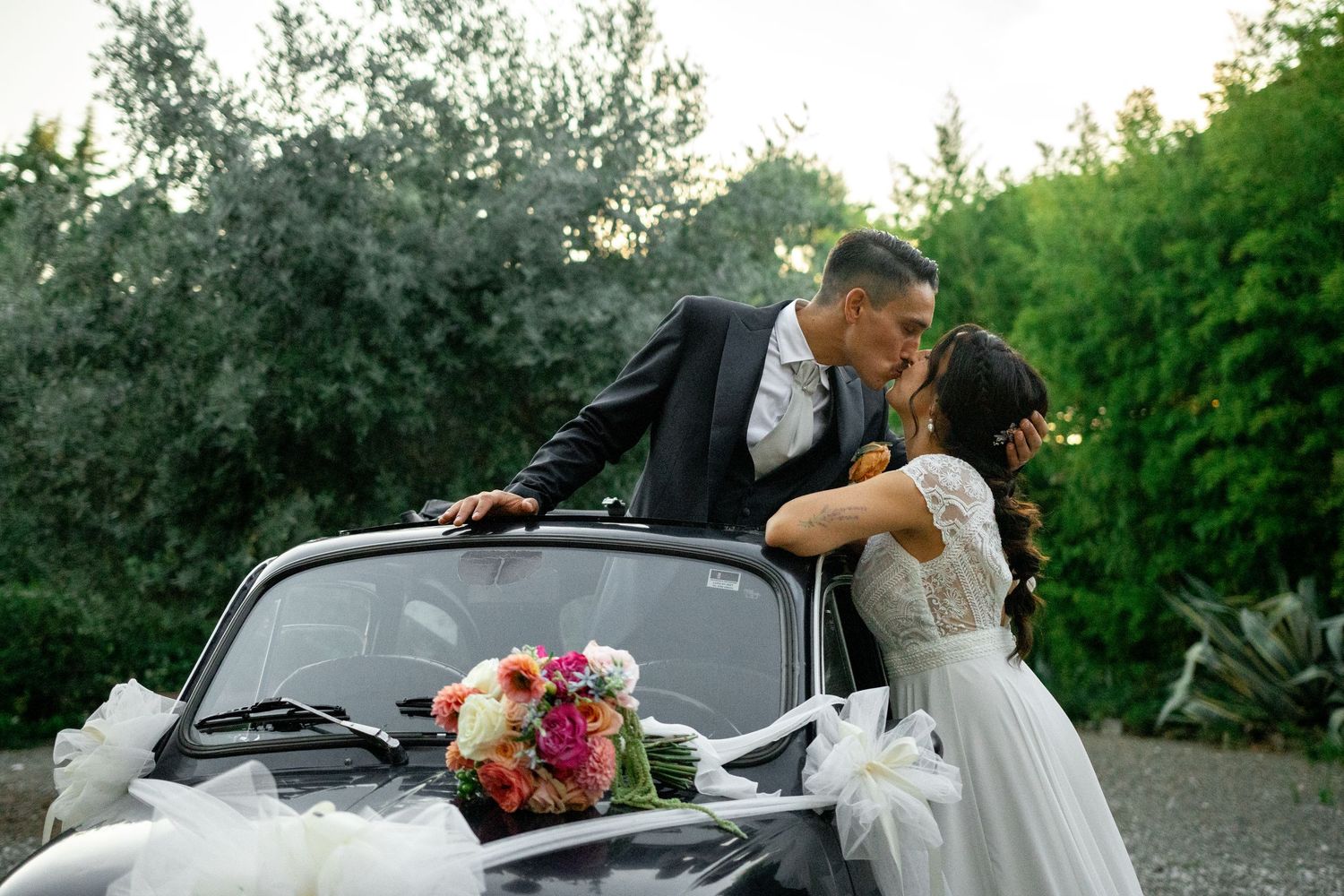 Bride and groom share a romantic kiss leaning over a vintage white car adorned with white ribbons and a colorful bouquet.