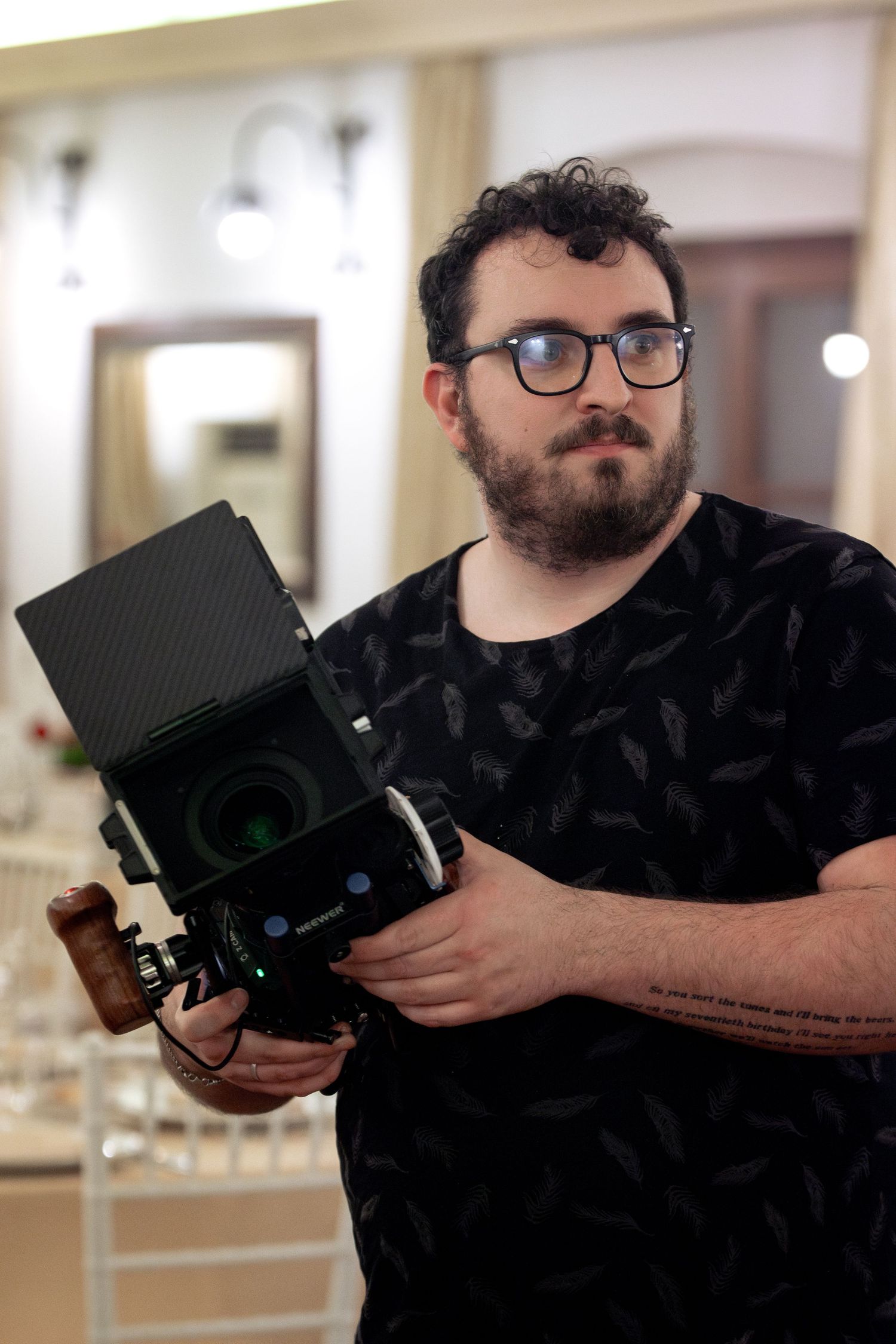 Bearded man with glasses holding a large professional camera in an indoor setting, wearing a black feather-print t-shirt.