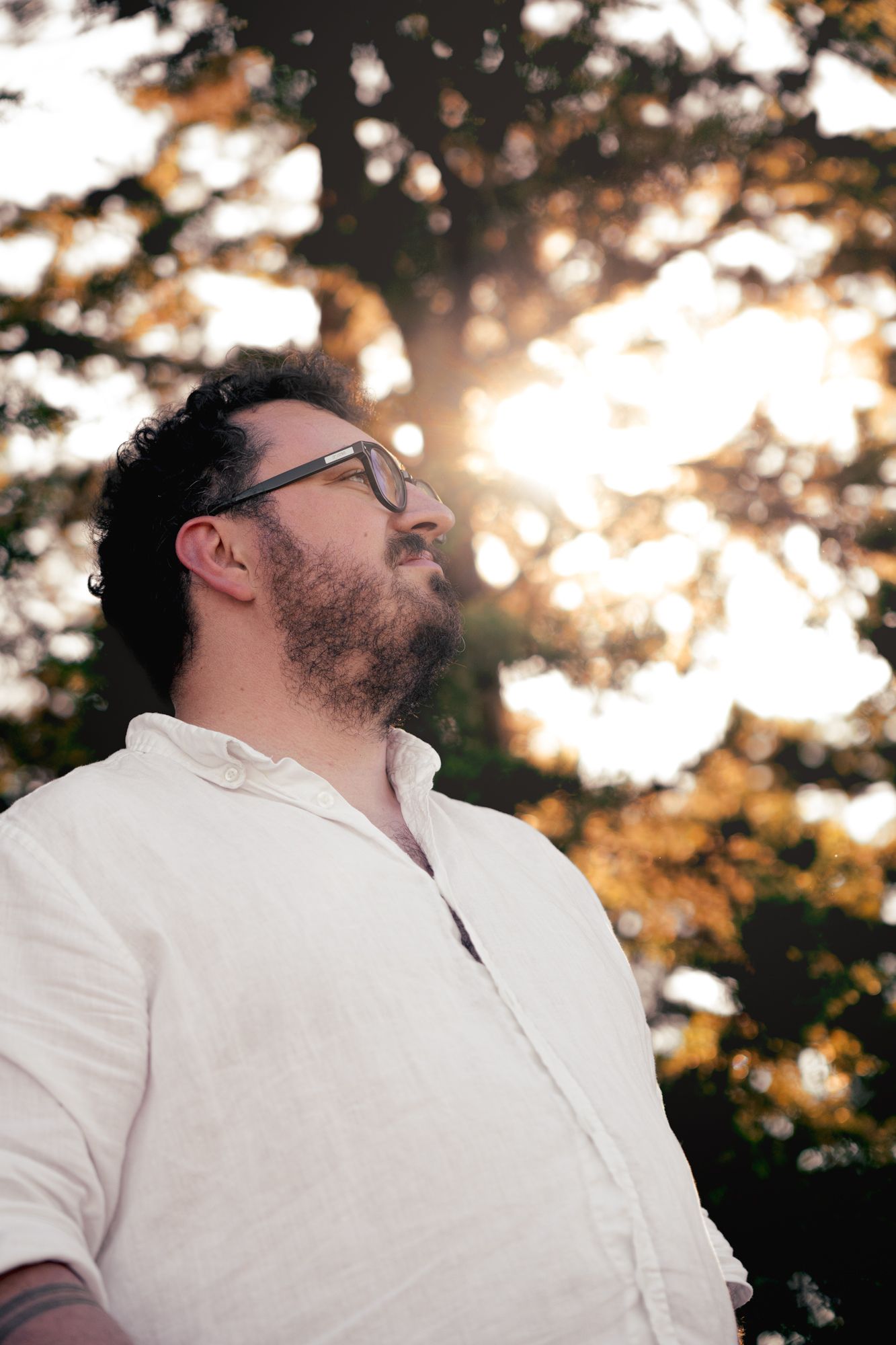 Bearded man with glasses in white linen shirt gazes upward outdoors, sun rays filtering through autumn trees.