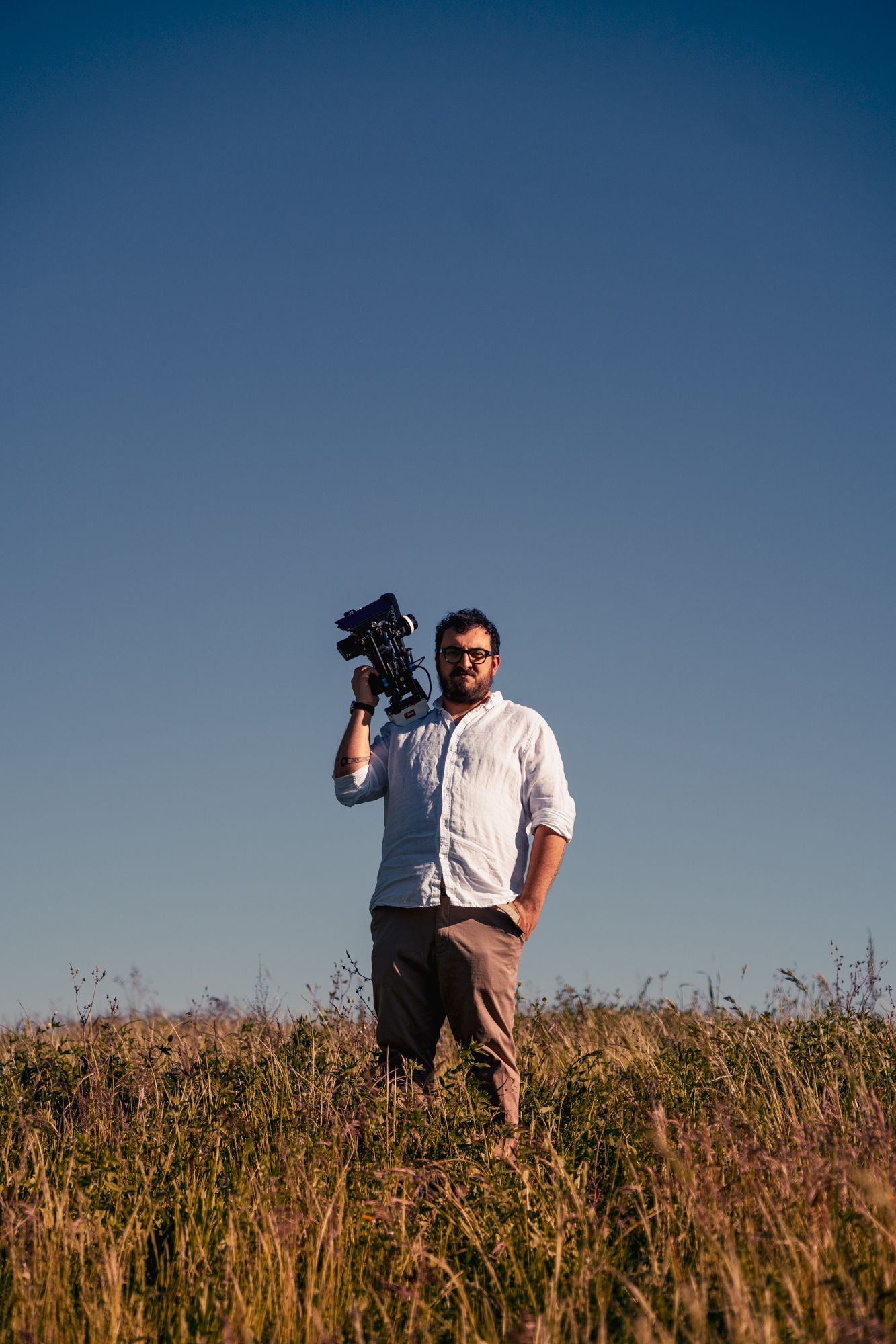 A bearded man in a white shirt holds a professional camera on his shoulder while standing in a golden field under a clear blue sky.
