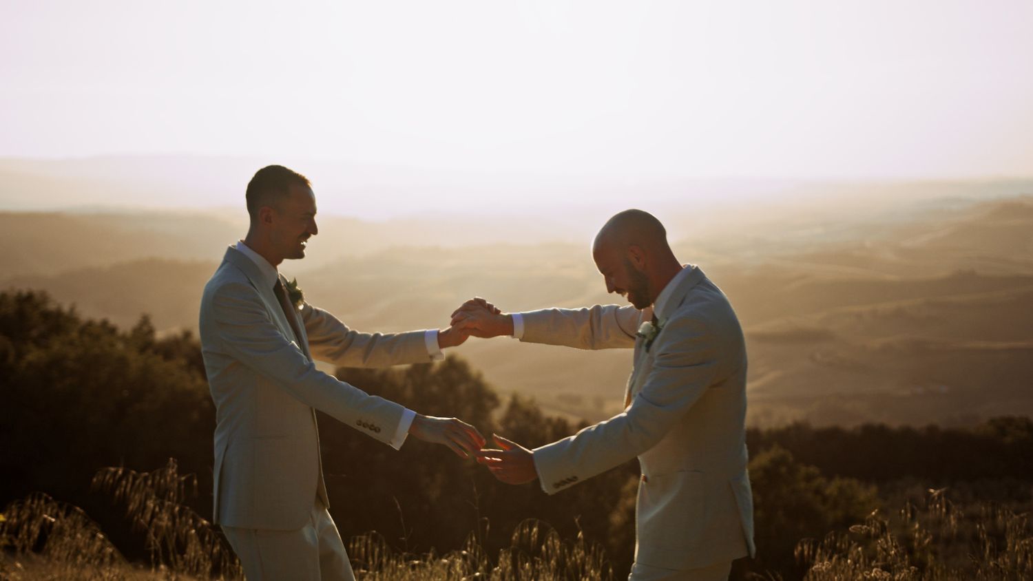Two grooms in light blue suits hold hands at sunset with rolling hills in the background at their outdoor wedding.