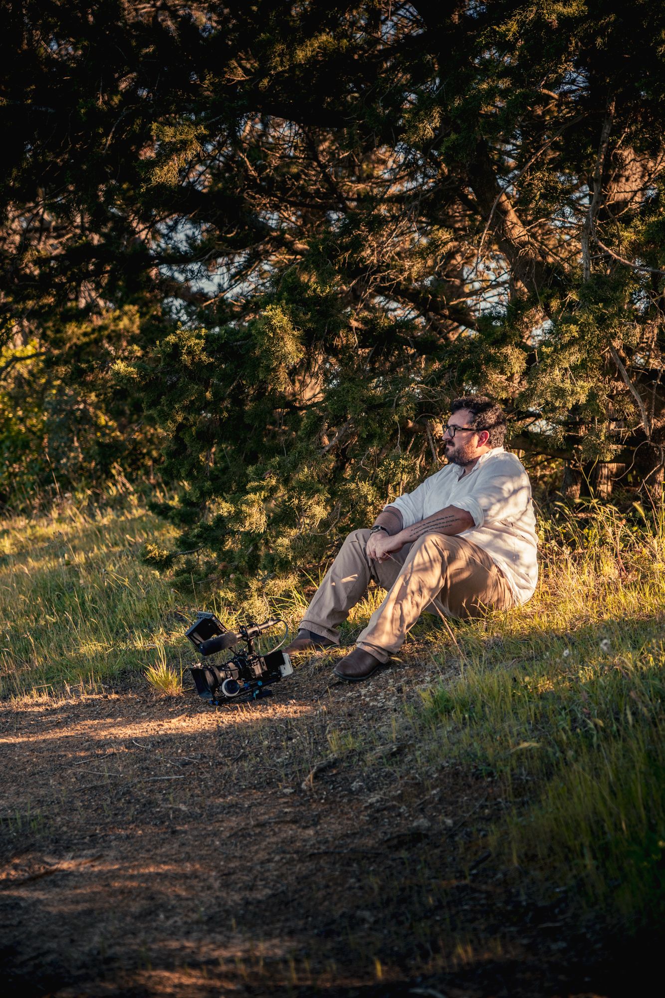 Man sitting under pine tree at golden hour with camera gear beside him on a dirt path.