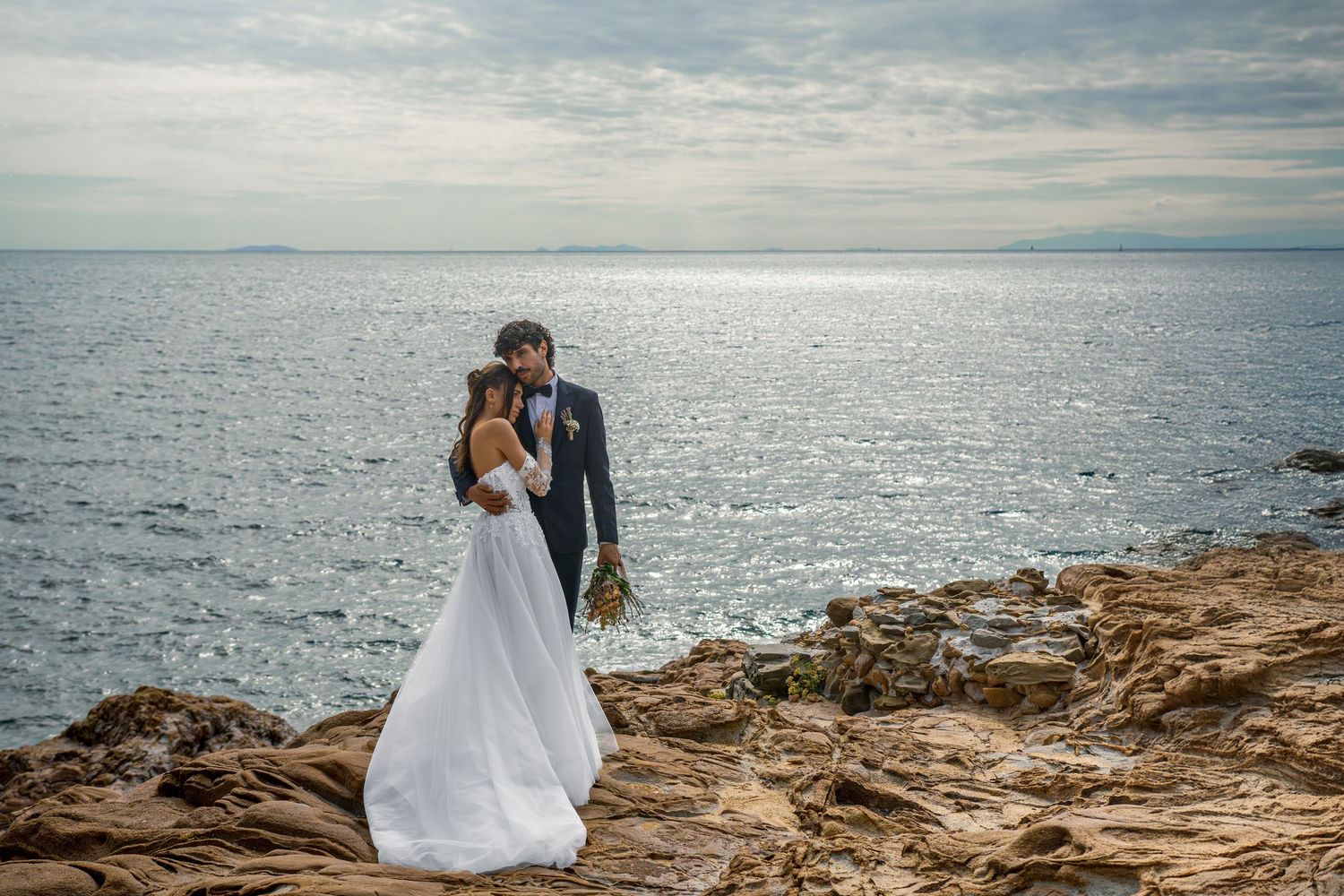 Bride and groom embrace on rocky coastal cliffs with shimmering ocean backdrop on their wedding day.