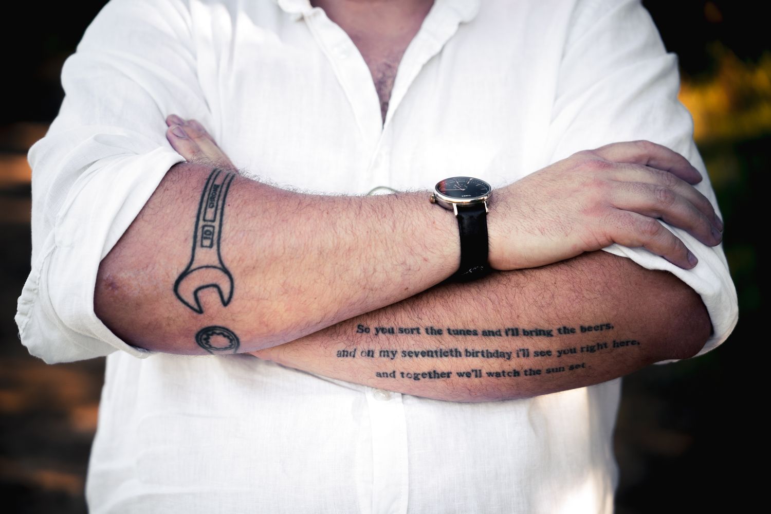 Man with crossed arms showing forearm tattoos of a wrench and cursive text quote, wearing a white shirt and watch.