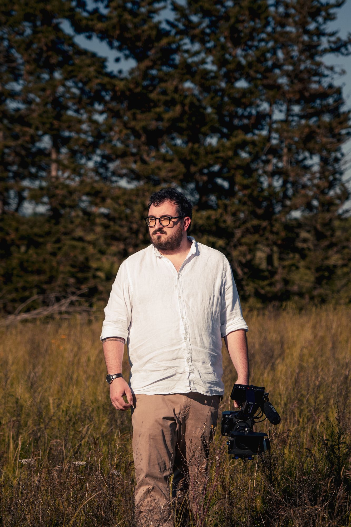 Bearded man with glasses holding a camera in a golden field with evergreen trees behind him.