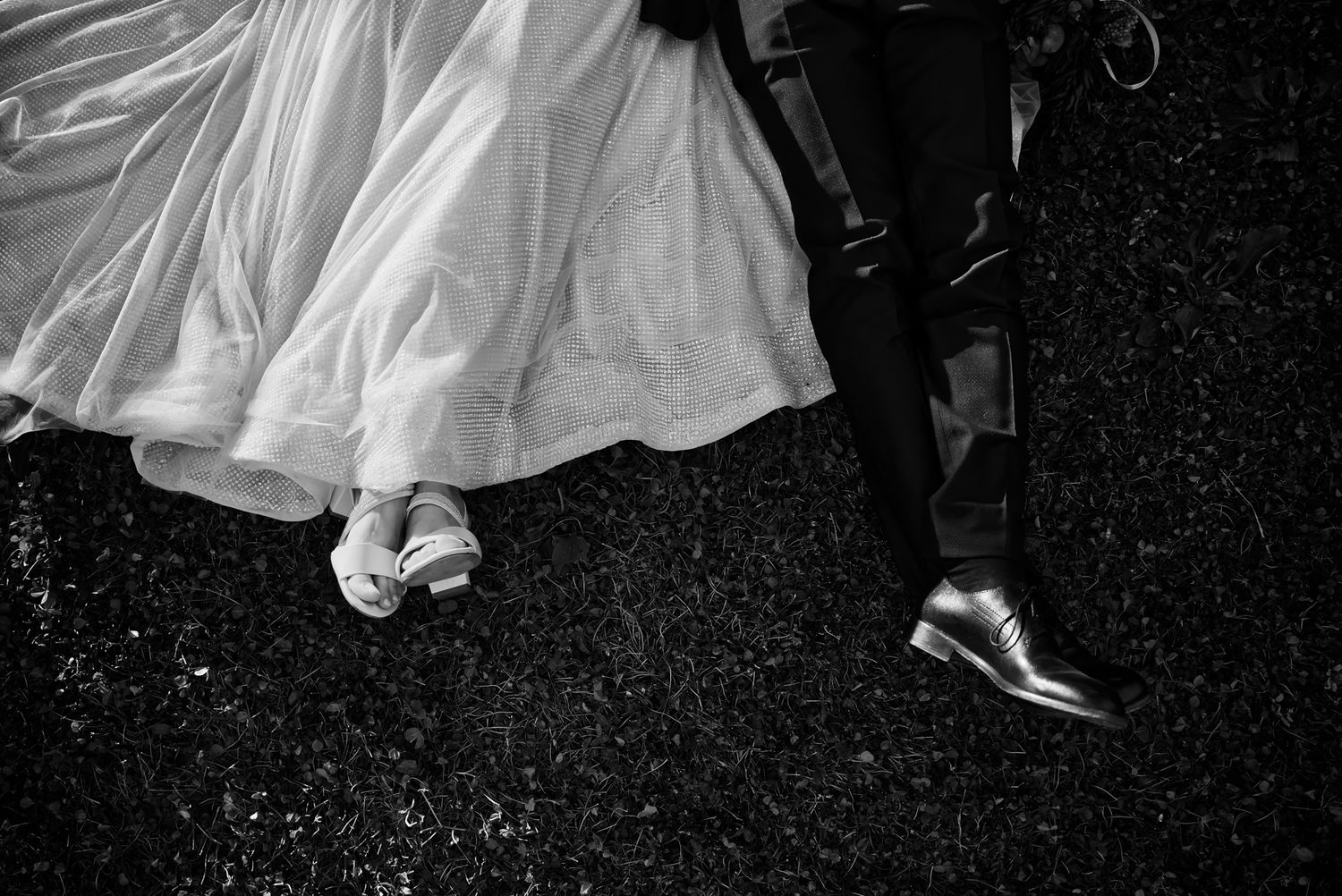 Black and white photo of bride and groom feet, showing white wedding dress hem and polished dress shoes on dark pavement.