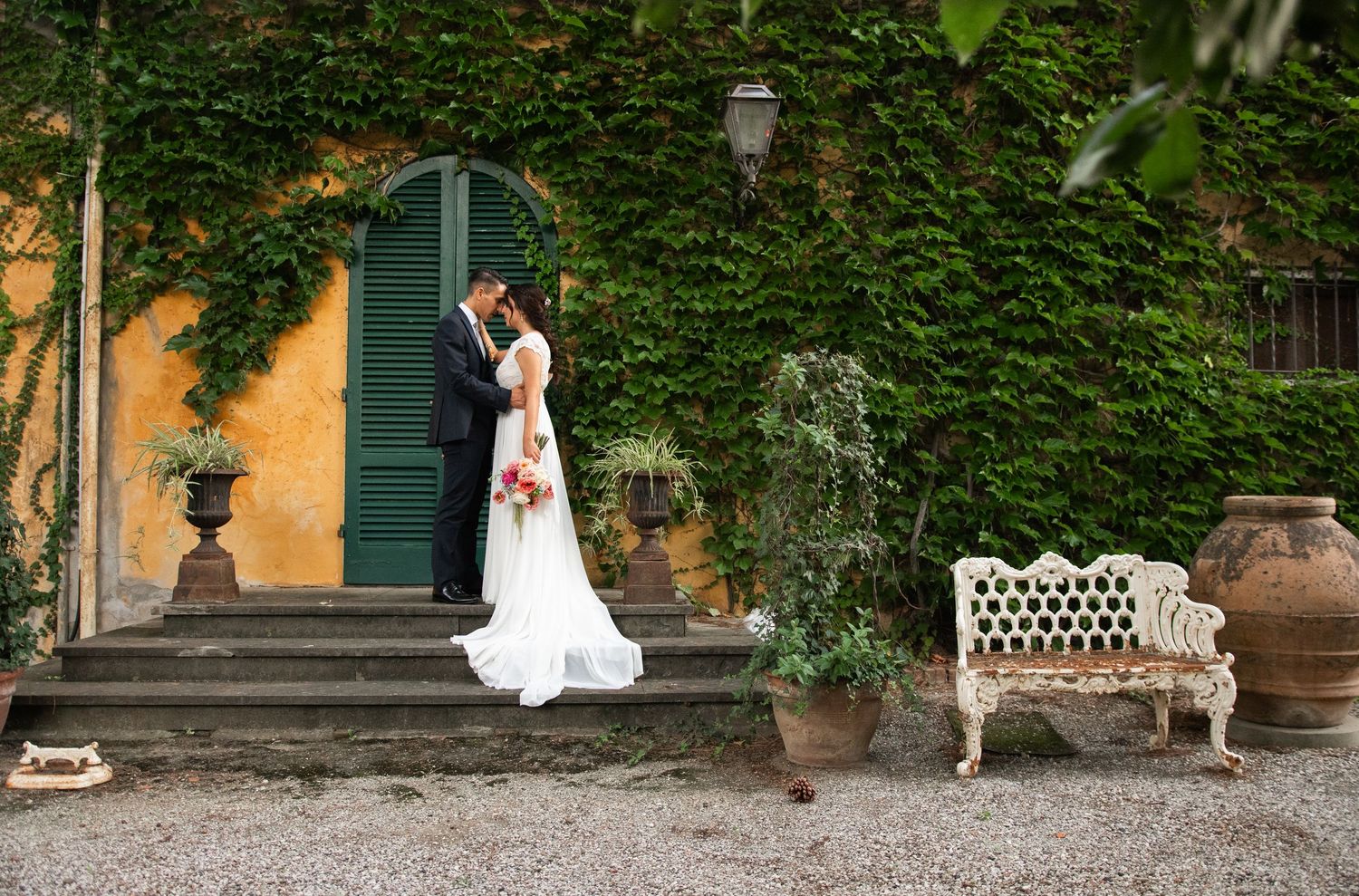 Bride and groom share a romantic kiss on stone steps of an ivy-covered villa with a white garden bench nearby.