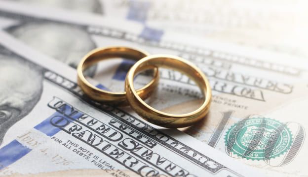 Two gold wedding rings resting on US hundred dollar bills, symbolizing marriage and financial planning.