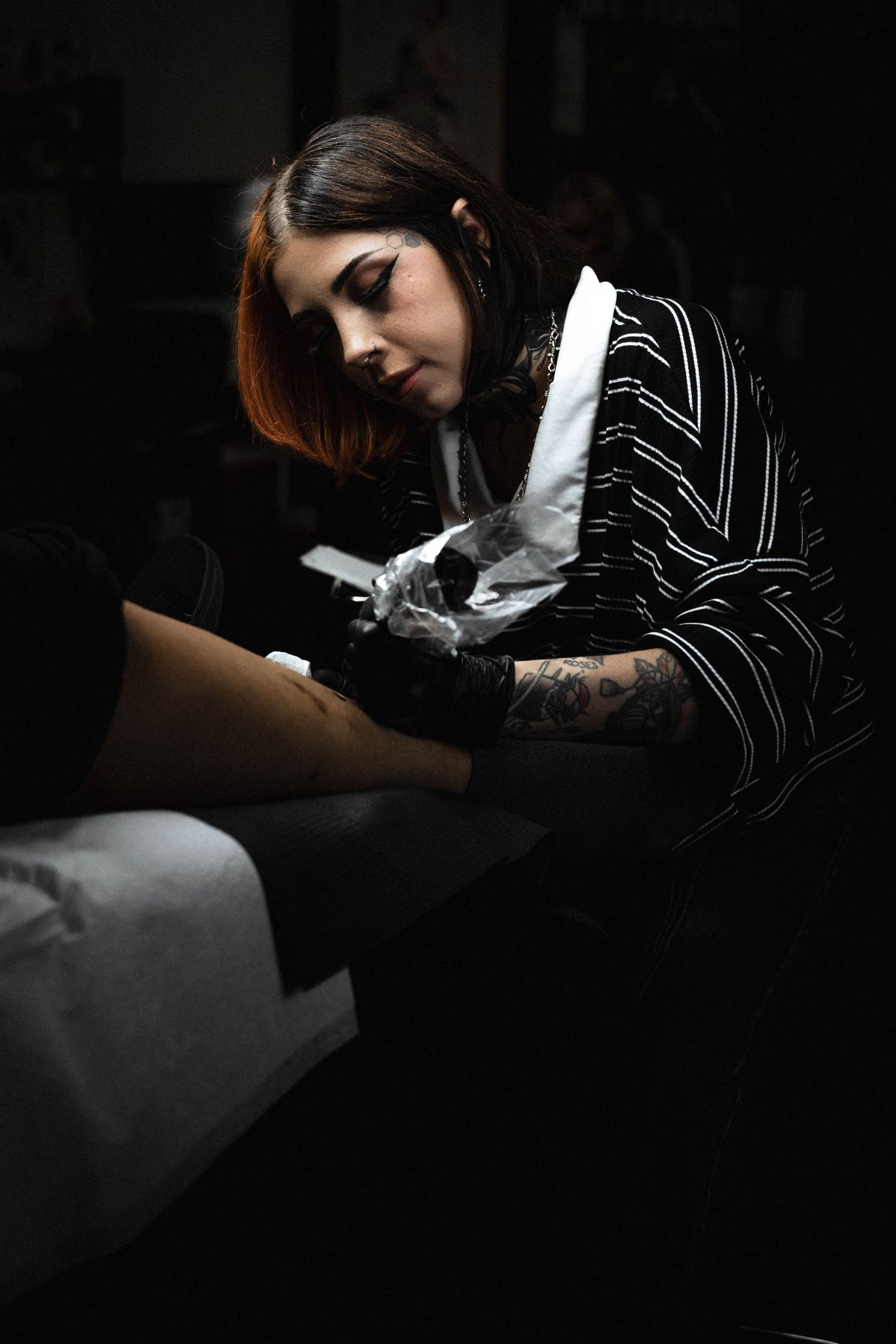 Tattoo artist wearing striped shirt works on client in dark moody studio setting with dramatic lighting.