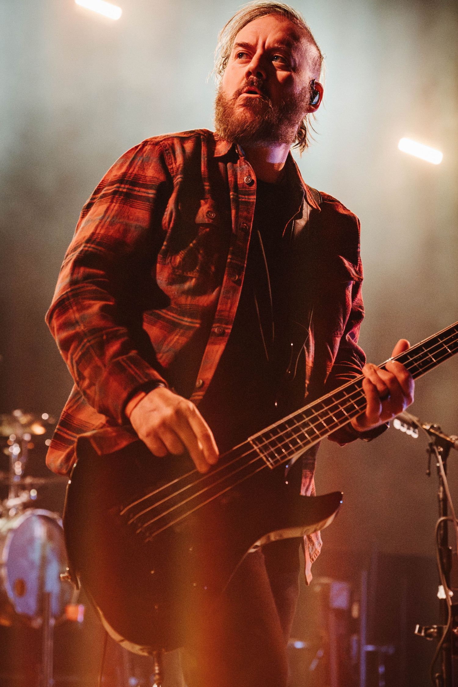 Musician in red plaid shirt plays bass guitar on dimly lit stage during live performance.