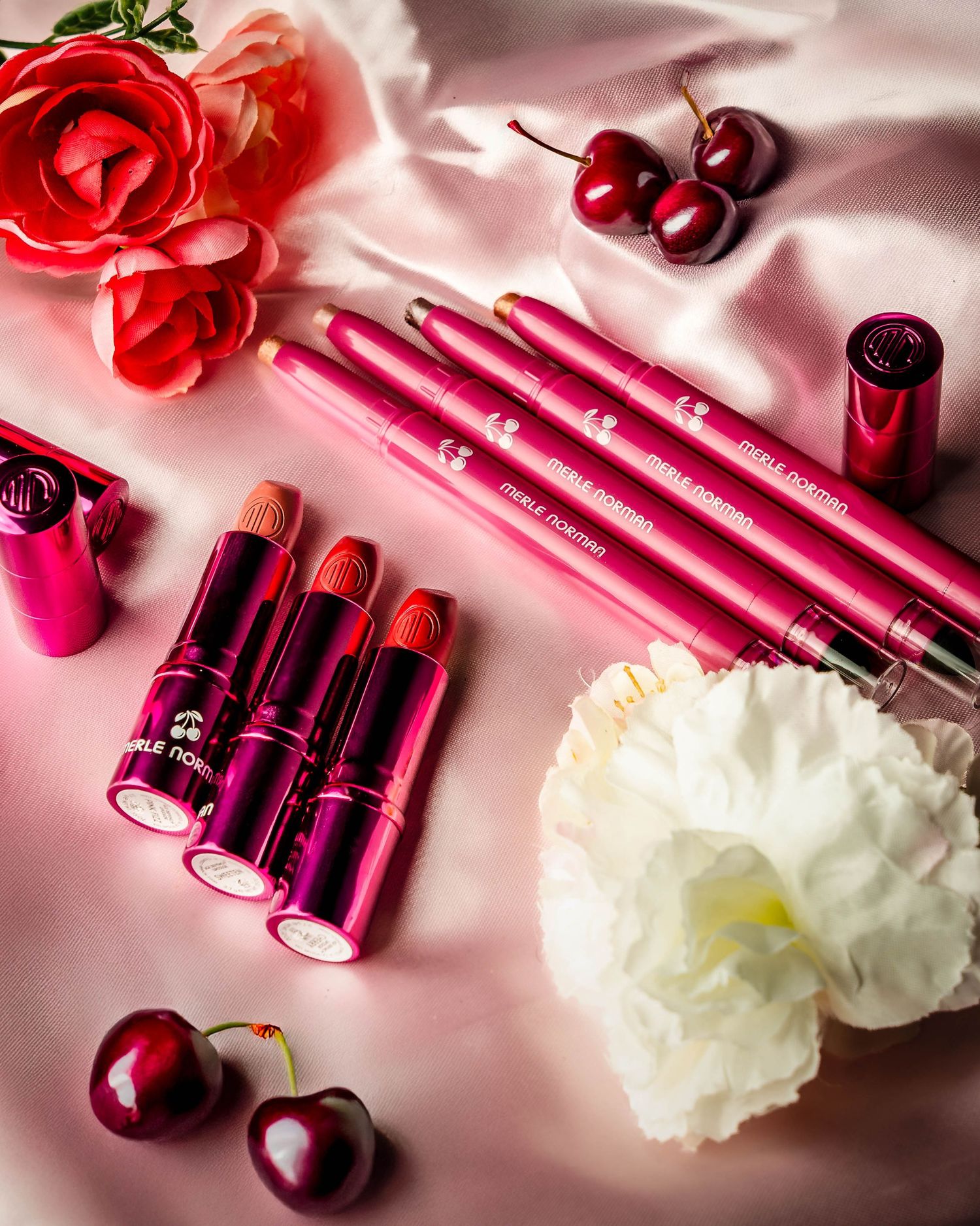 Flatlay of pink makeup products with roses and cherries on pink silk fabric.