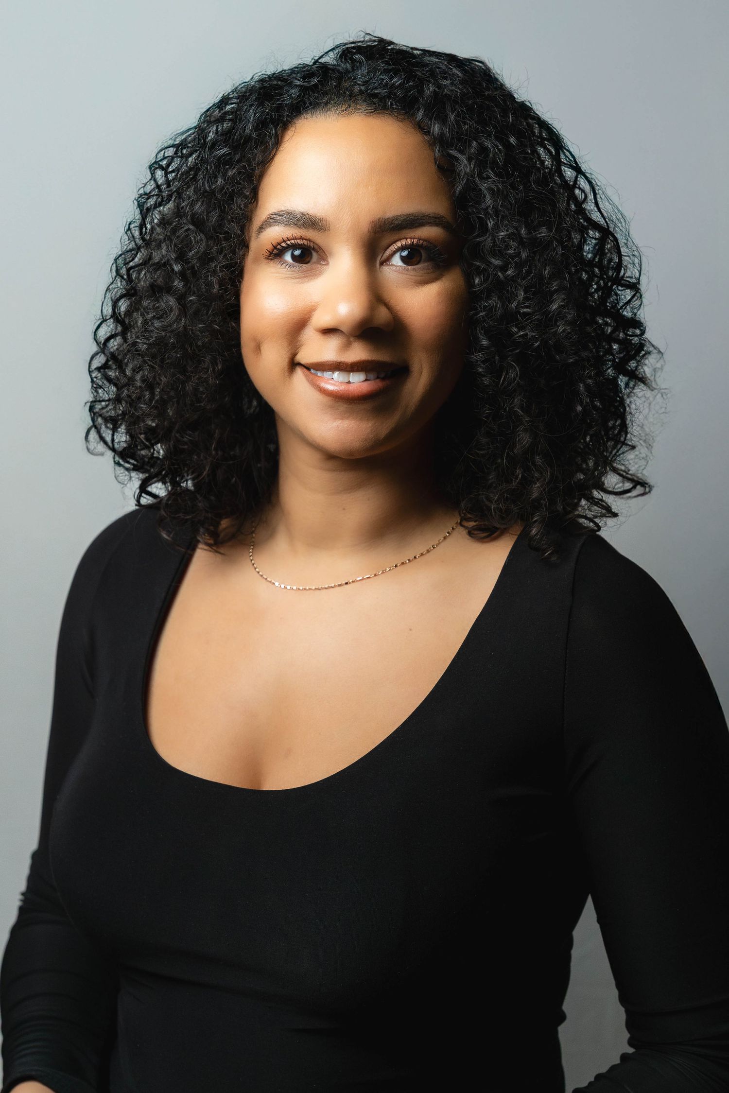Professional portrait of a person with natural curly hair wearing a black top and delicate gold necklace against light background.
