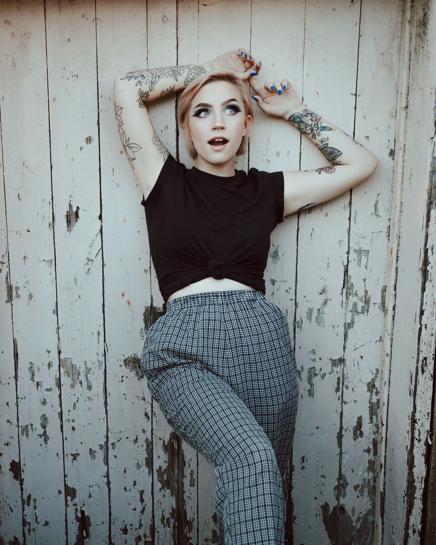 Alternative fashion model in black crop top and plaid pants poses against weathered wooden wall.