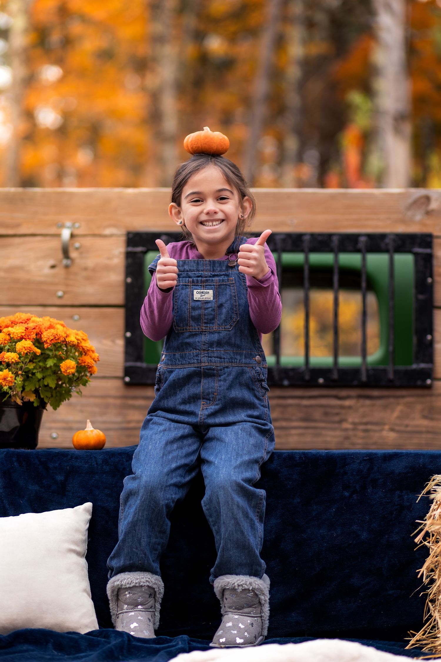 A smiling girl in denim overalls balances a mini pumpkin on her head giving thumbs up in an autumn outdoor setting.
