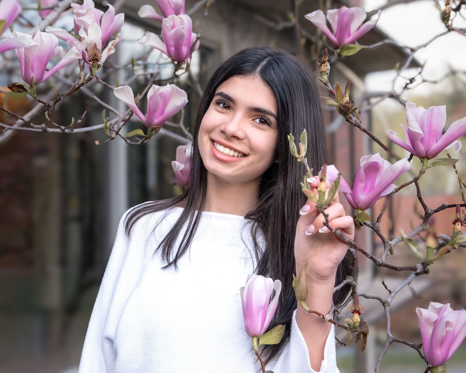 Young woman smiling among pink magnolia blossoms in spring, wearing a white sweater outdoors.