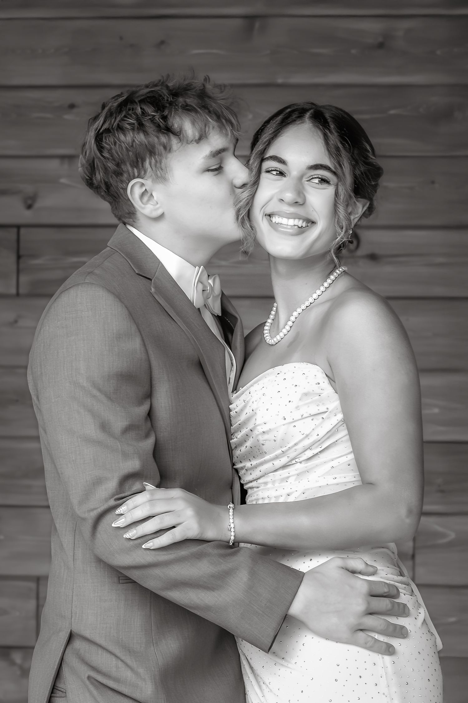 Black and white photo of a couple in formal wear, groom kissing bride on cheek as she smiles joyfully.
