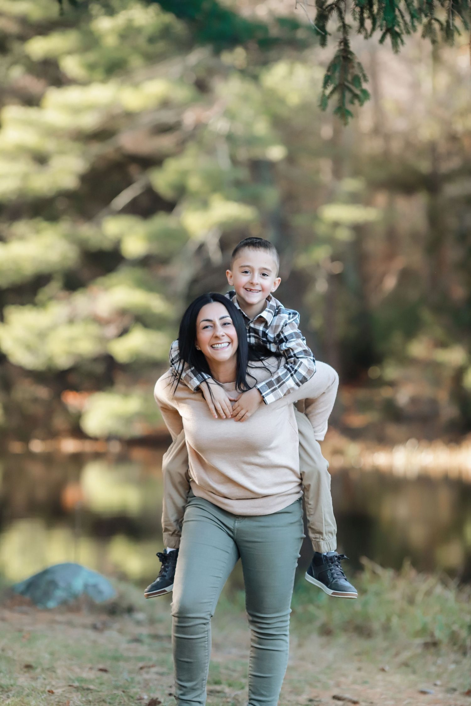 A smiling woman gives a happy young boy a piggyback ride outdoors near a forest lake in autumn.