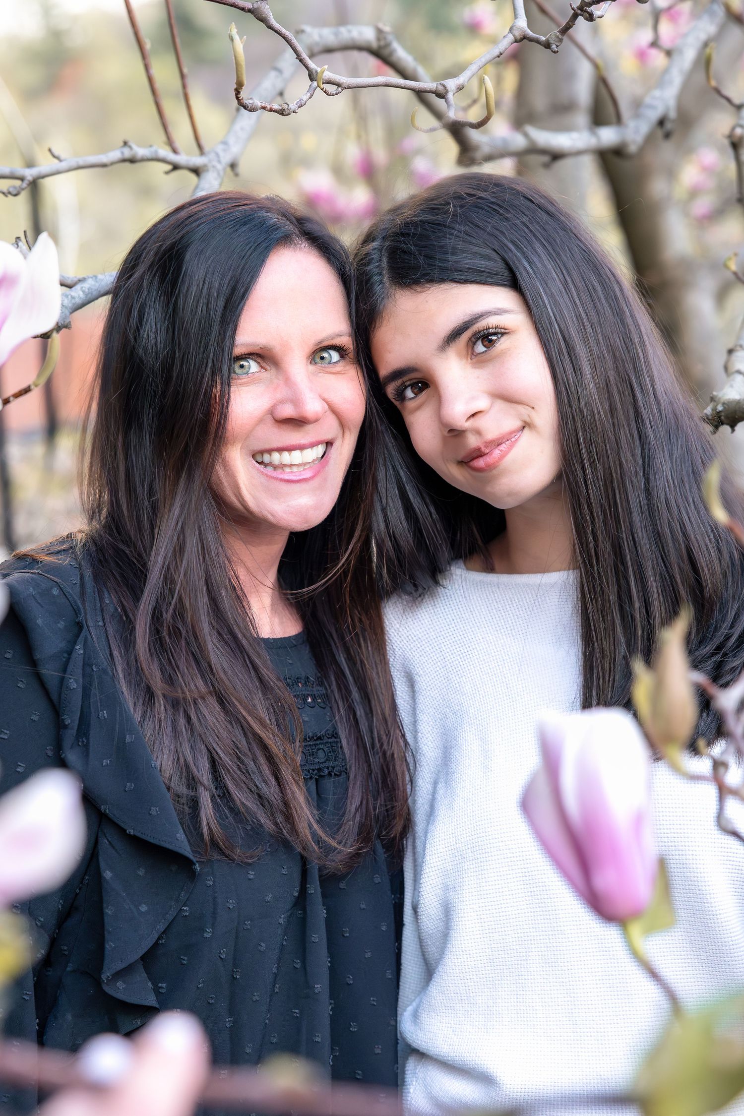 Two women with dark hair smile together outdoors among pink magnolia blossoms in spring.