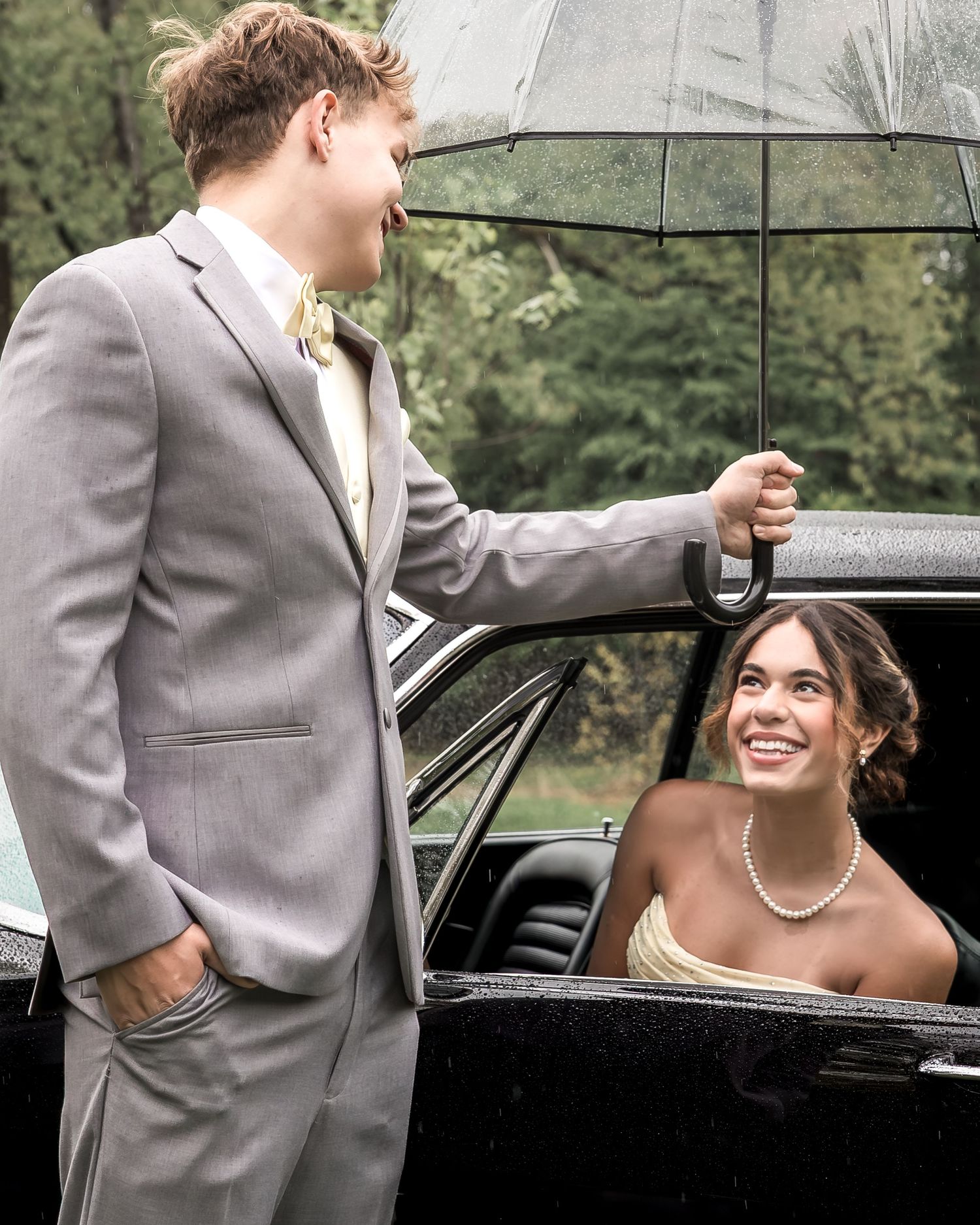 Young man in gray suit holds umbrella for smiling woman in formal dress seated in black car on rainy day.